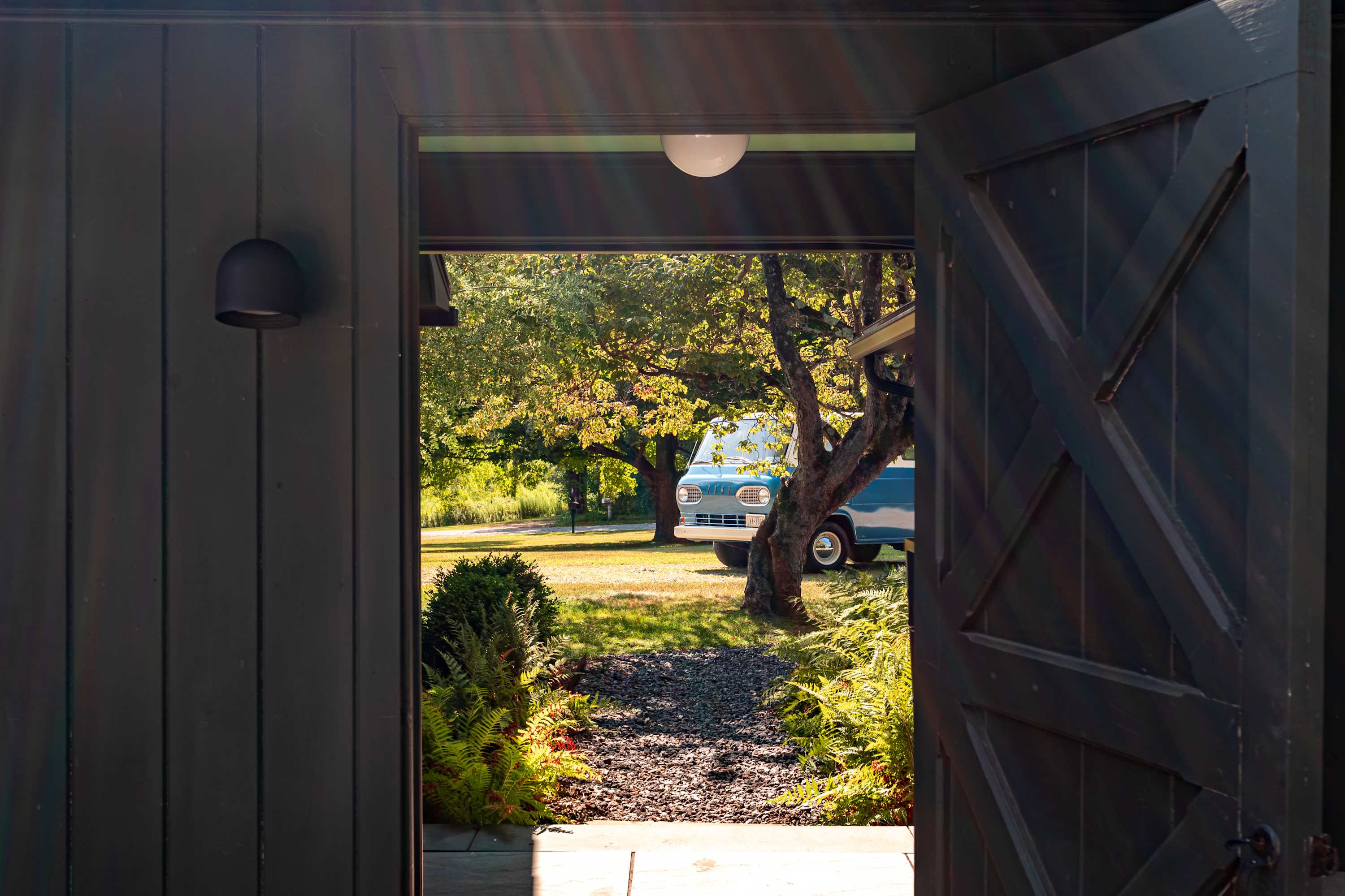 View through an open dark wooden doorway, revealing a gravel path leading to a sunny green yard with mature trees and a vintage blue van parked under them. Sunlight streams through the top of the doorway.