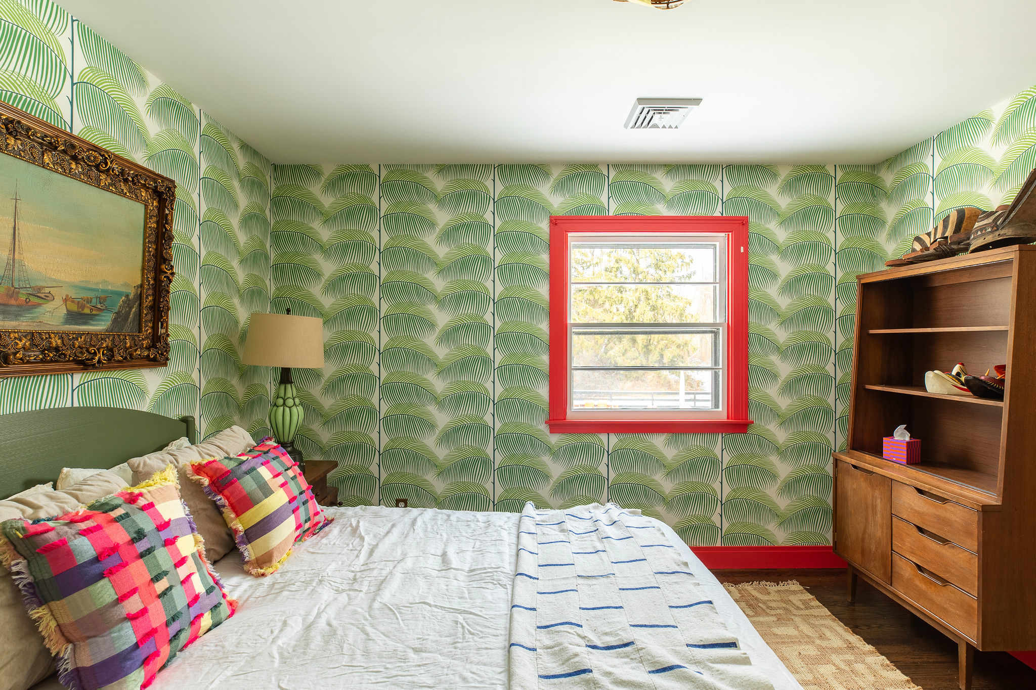 Bedroom with striking green palm leaf wallpaper, showcasing a window framed in red trim, a wooden dresser with shelves, and a bed with white bedding. A woven rug is visible on the floor.