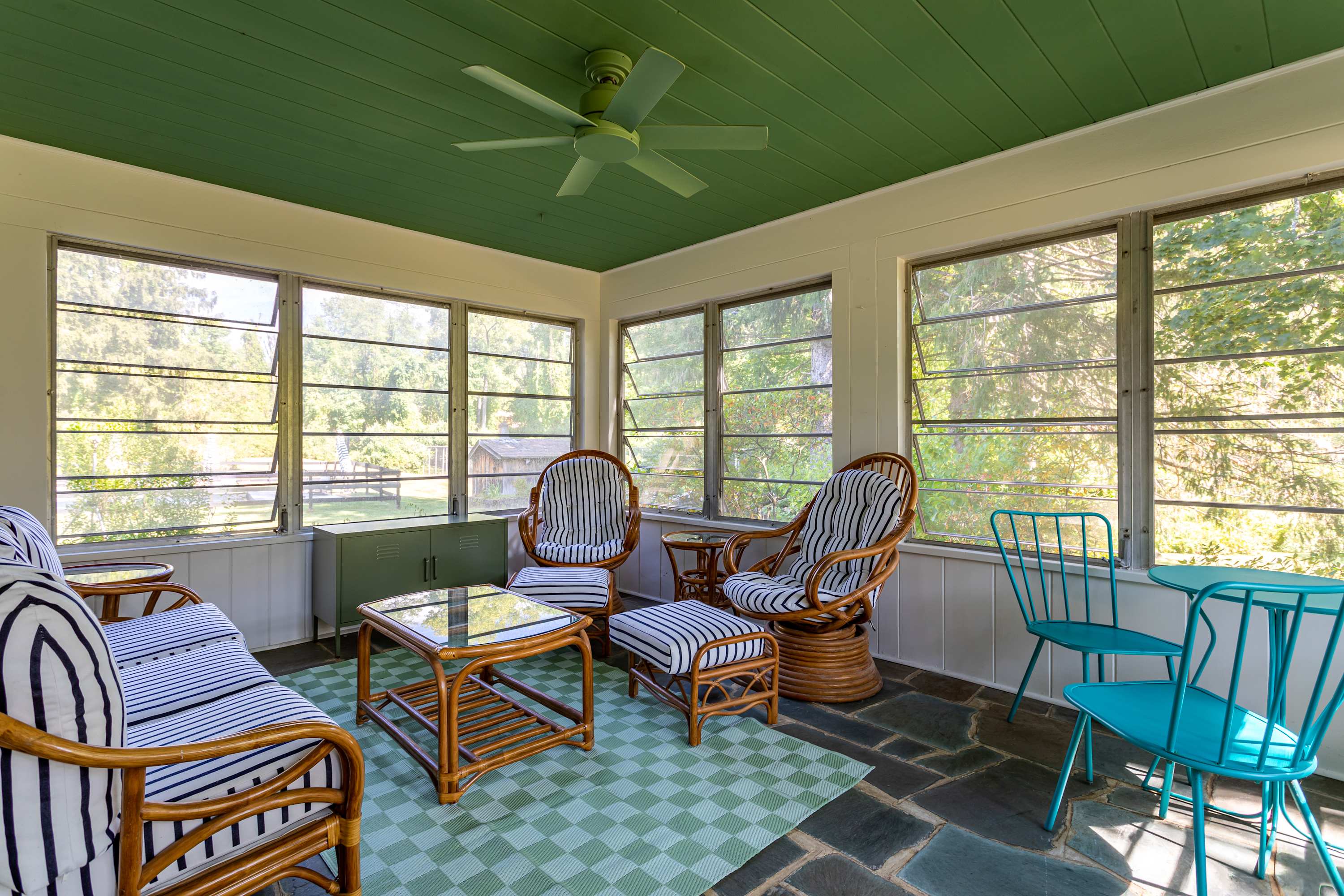 A spacious screened porch with a vibrant green slatted ceiling, white paneled walls, and a dark slate floor. It's furnished with rattan seating, a glass-top coffee table, a small blue bistro set, and a green and white checkered rug.