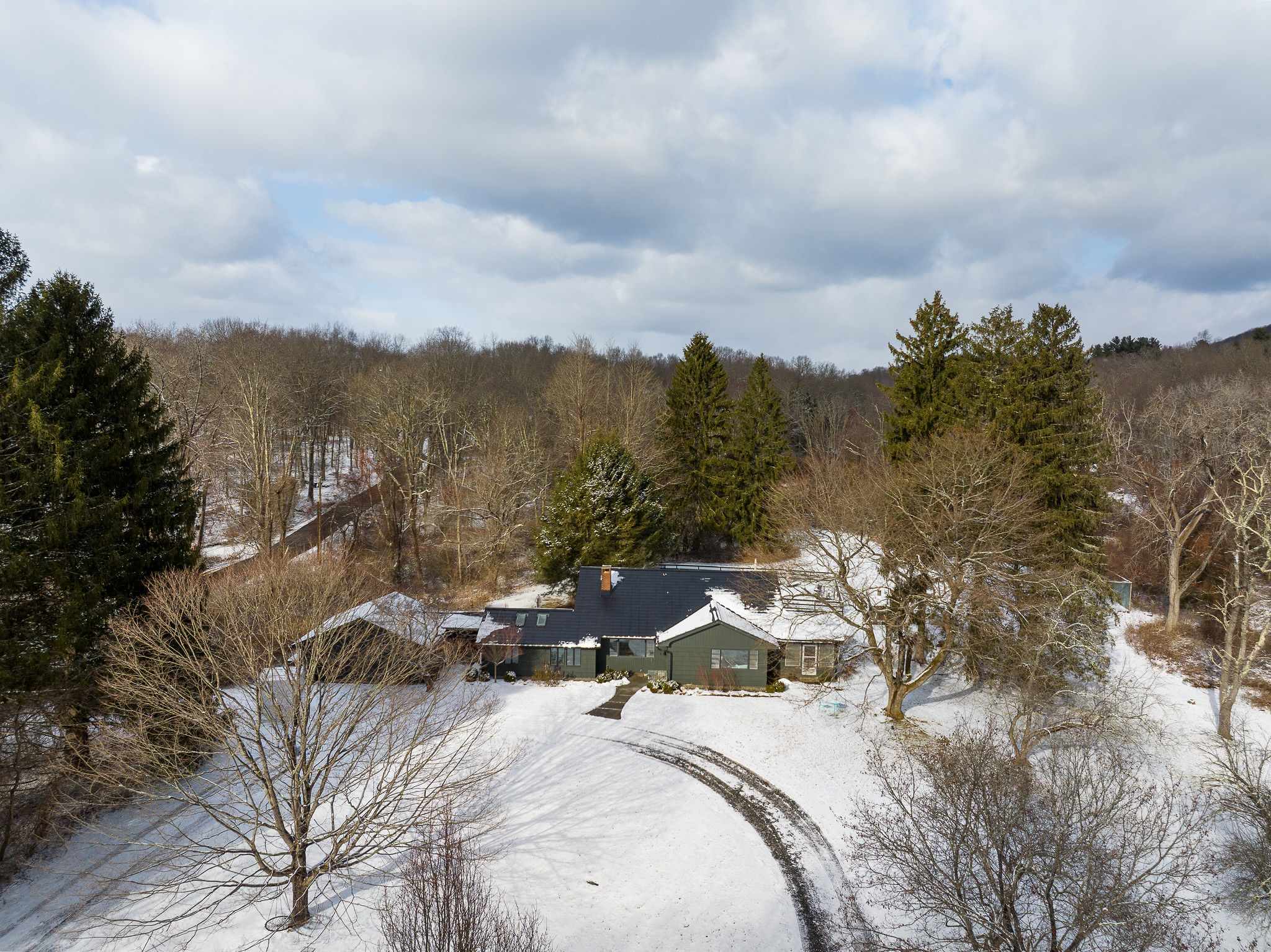 An aerial view of the dark gray house nestled among snow-covered trees and a winding driveway, showcasing its secluded location in a winter landscape. The roof is dark, and the surrounding forest is bare.