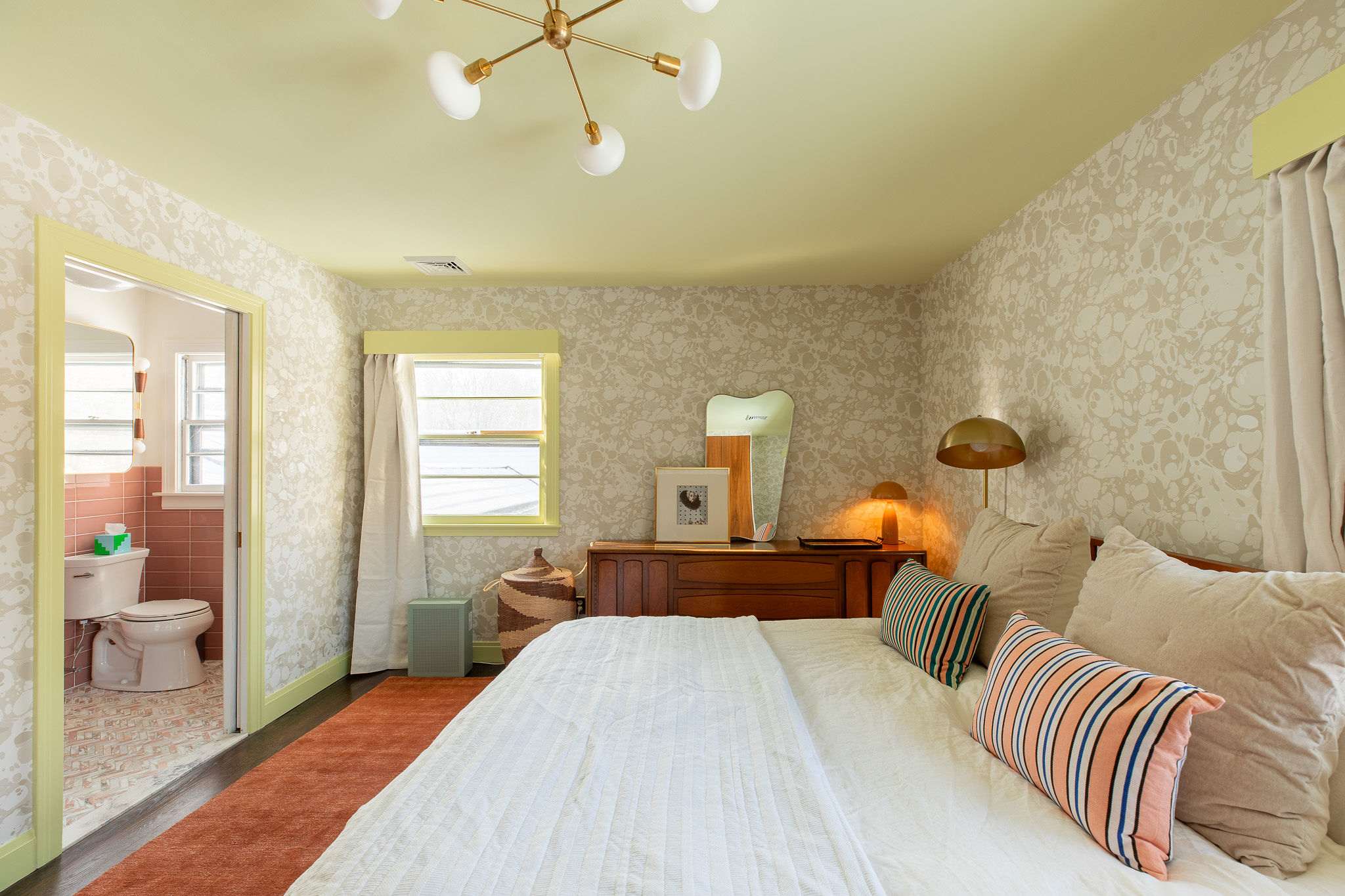 View of the bedroom showing the wooden bed with white bedding and striped pillows, a mid-century dresser with a mirror, and a doorway leading to a pink-tiled bathroom. Light green trim frames the windows and doorway.
