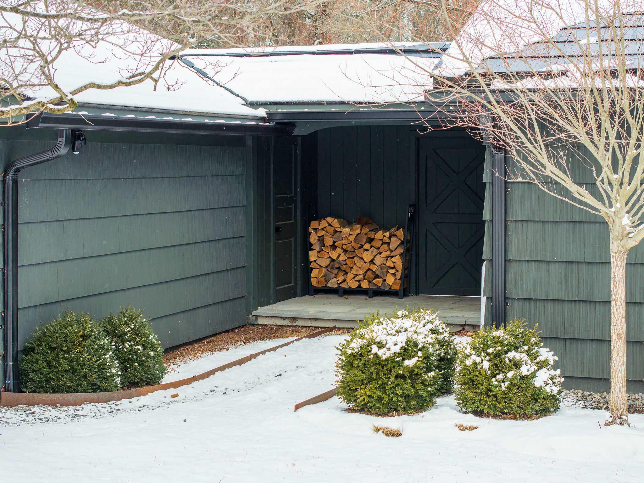 A covered outdoor area of the dark gray house, featuring a neatly stacked pile of firewood on a metal rack, ready for use. The ground is covered in snow, with small evergreen bushes and a bare tree nearby.