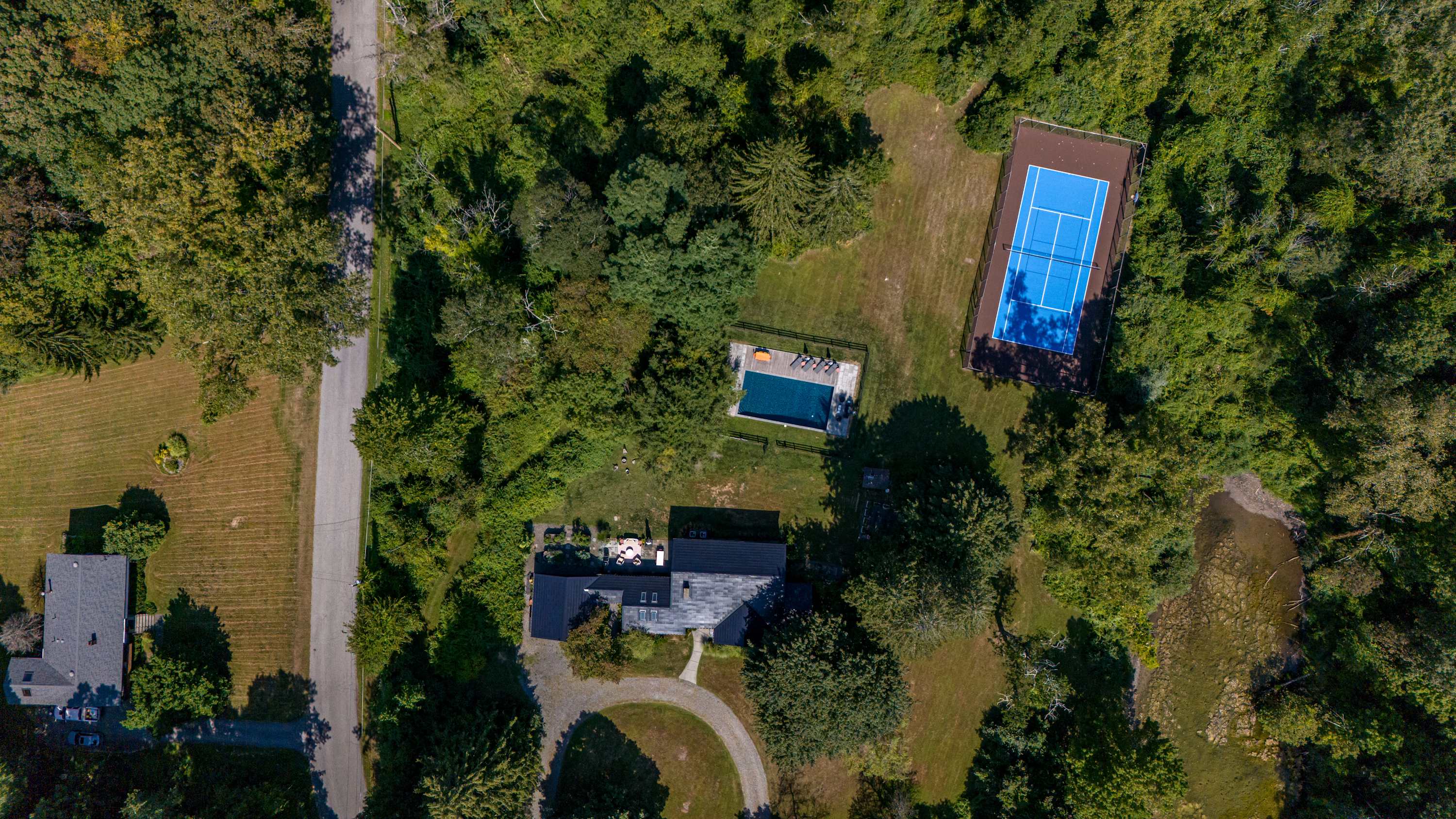 An aerial view of a large property featuring a dark-roofed house, a rectangular swimming pool, and a blue tennis court, all surrounded by dense green trees and a paved road on the left.