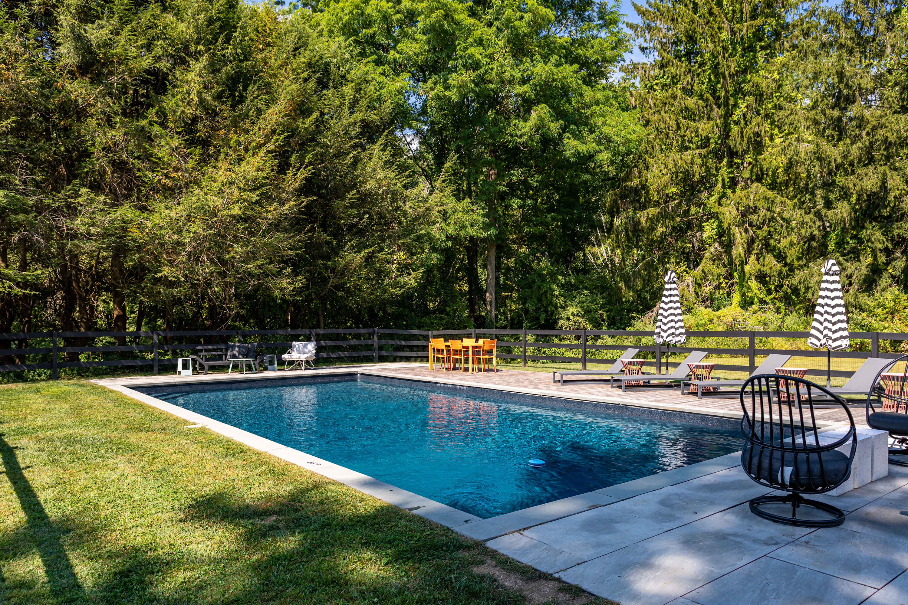 A rectangular inground swimming pool with dark blue water, surrounded by a concrete deck and a grassy lawn, featuring several lounge chairs and striped umbrellas, all enclosed by a black fence and mature trees.