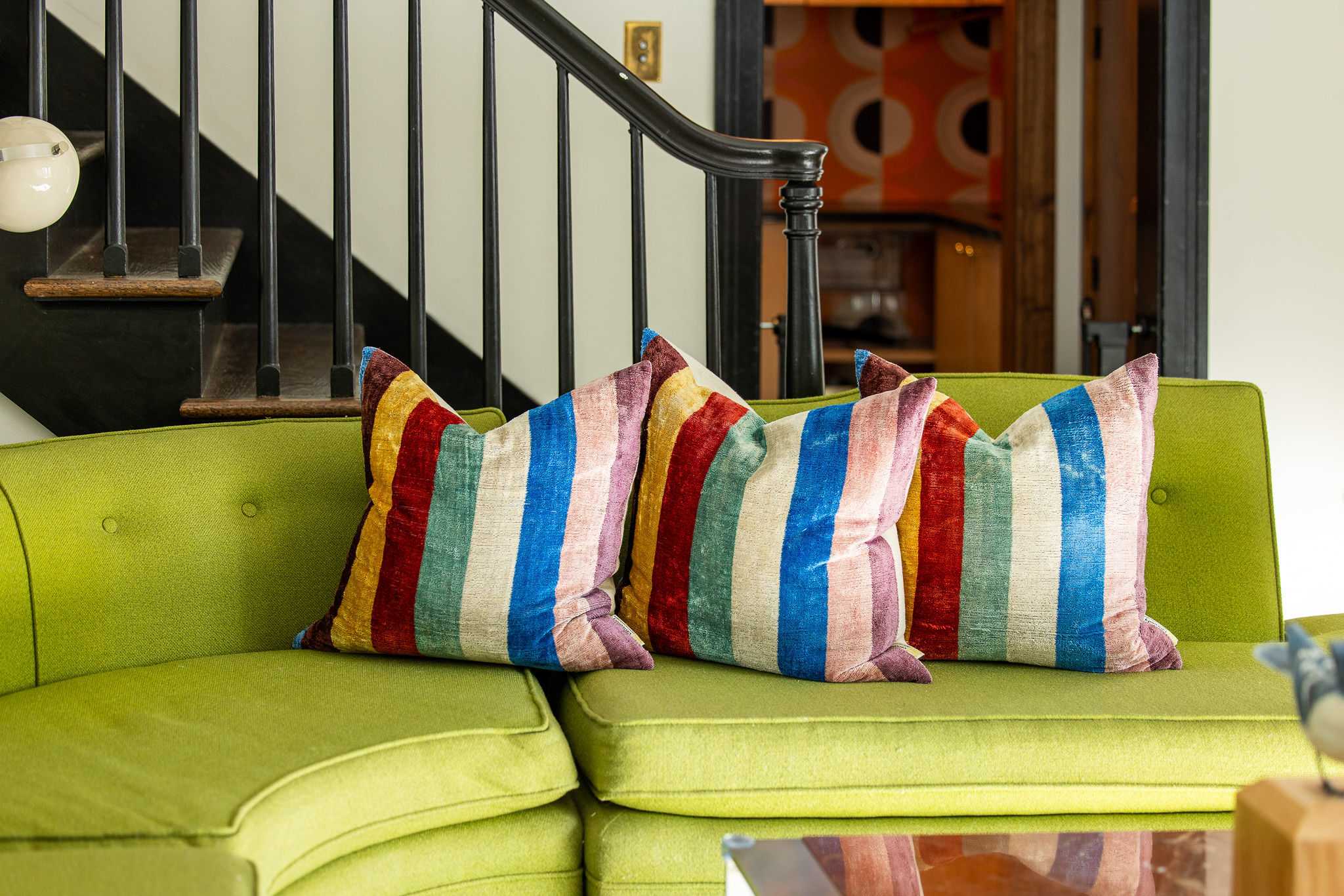 A close-up of a lime green sectional sofa adorned with three plush throw pillows featuring bold, multi-colored vertical stripes, set against a dark staircase railing.