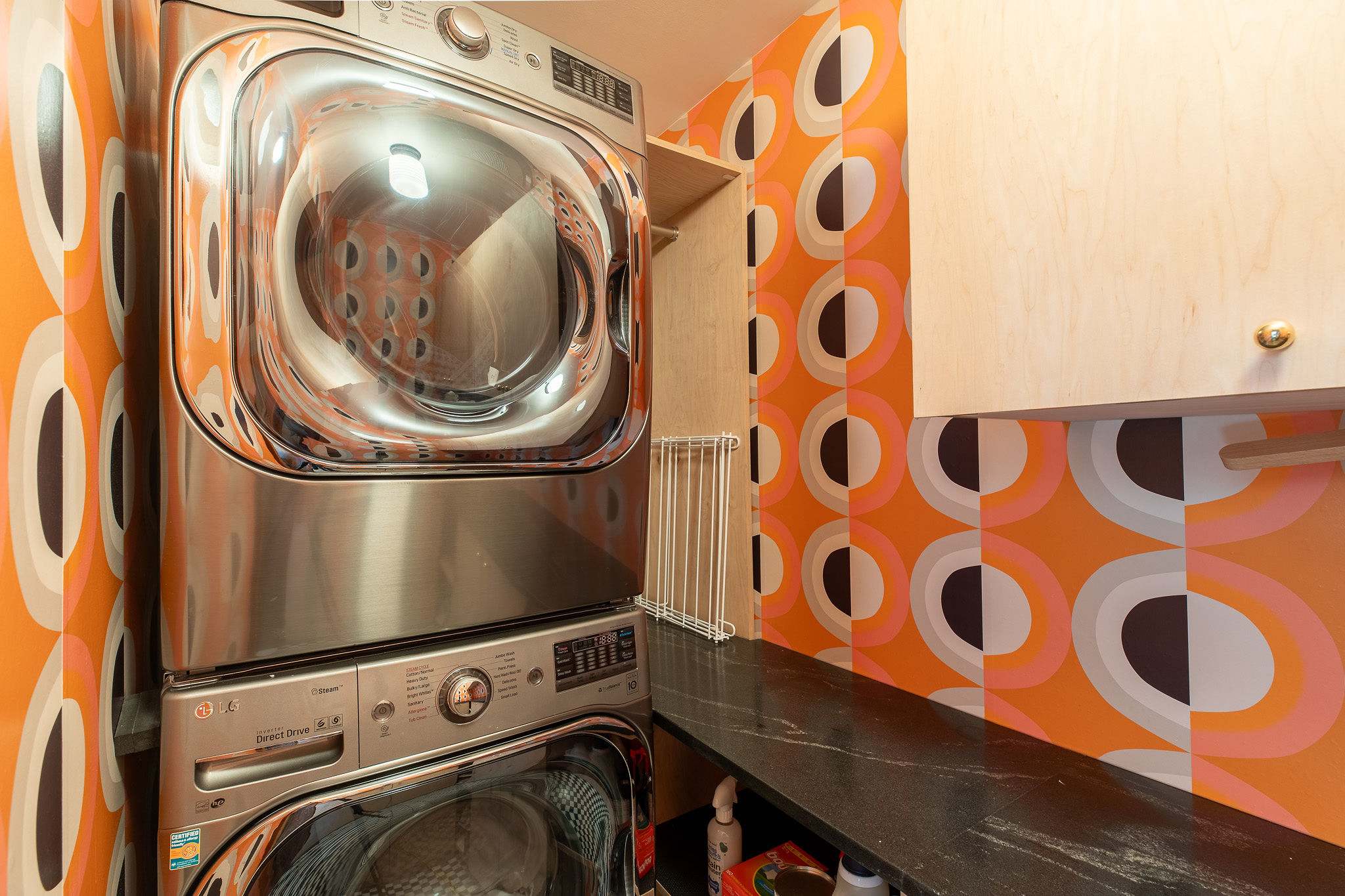 A modern laundry area featuring stacked stainless steel washer and dryer units, set against a bold orange and black geometric patterned wallpaper. A dark countertop and light wood shelving provide functional space.