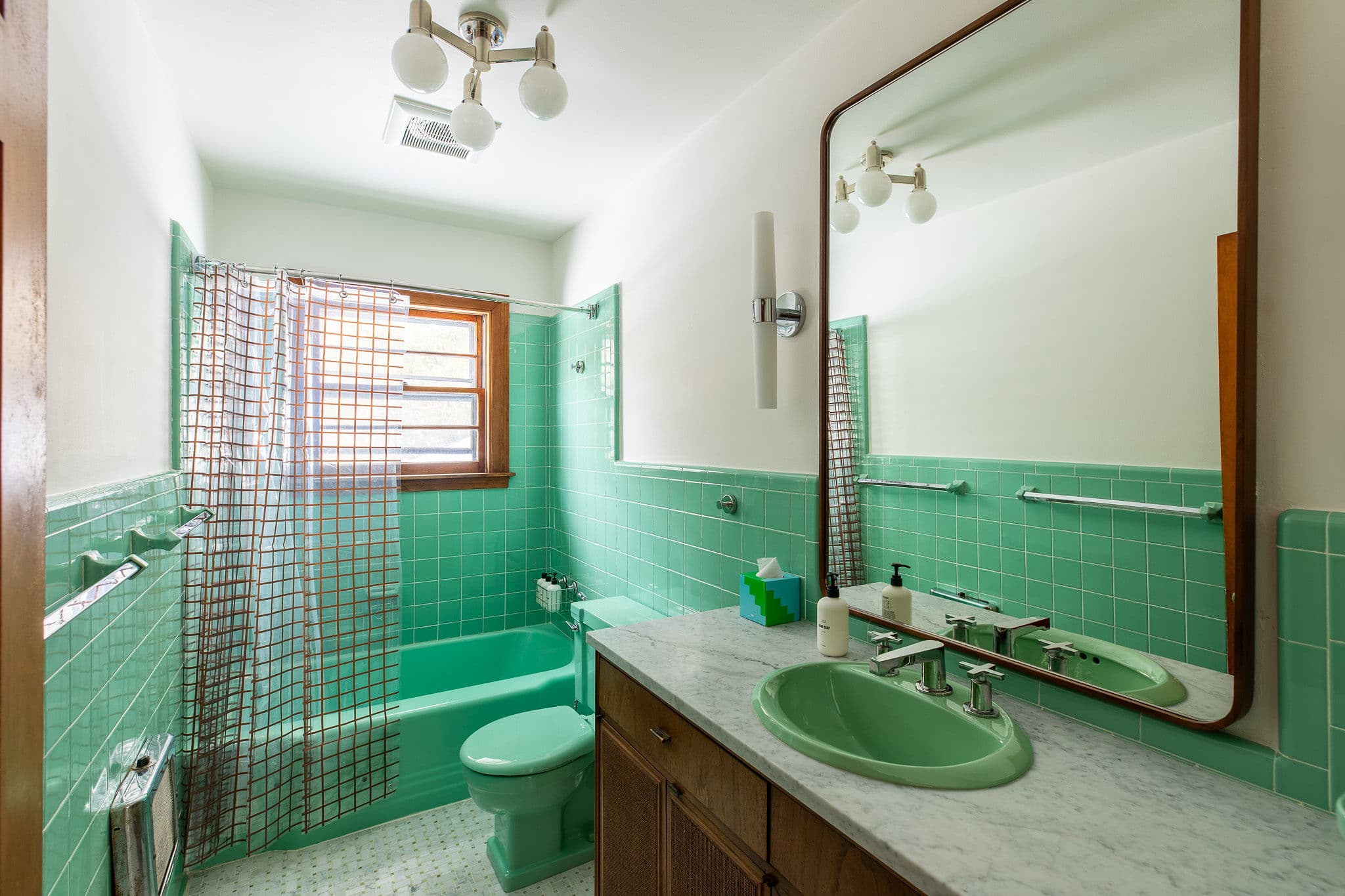 A retro-style bathroom featuring bright mint green subway tiles on the walls and floor, a matching green bathtub, toilet, and sink, complemented by a white marble vanity and a large mirror.
