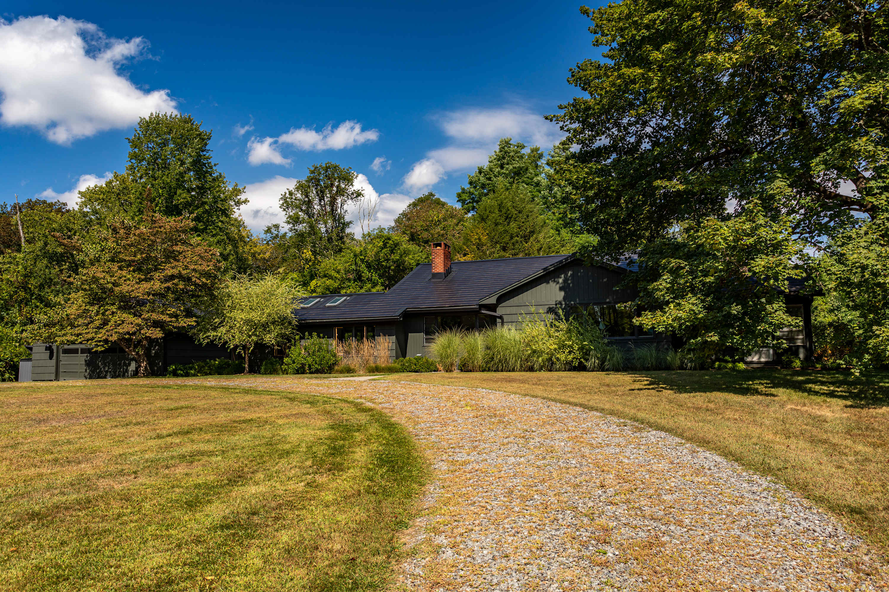 A wide shot of a dark-colored, single-story house with a dark roof and a brick chimney, nestled among abundant green trees. A gravel driveway leads up to the house, with a well-maintained lawn in the foreground under a bright blue sky.