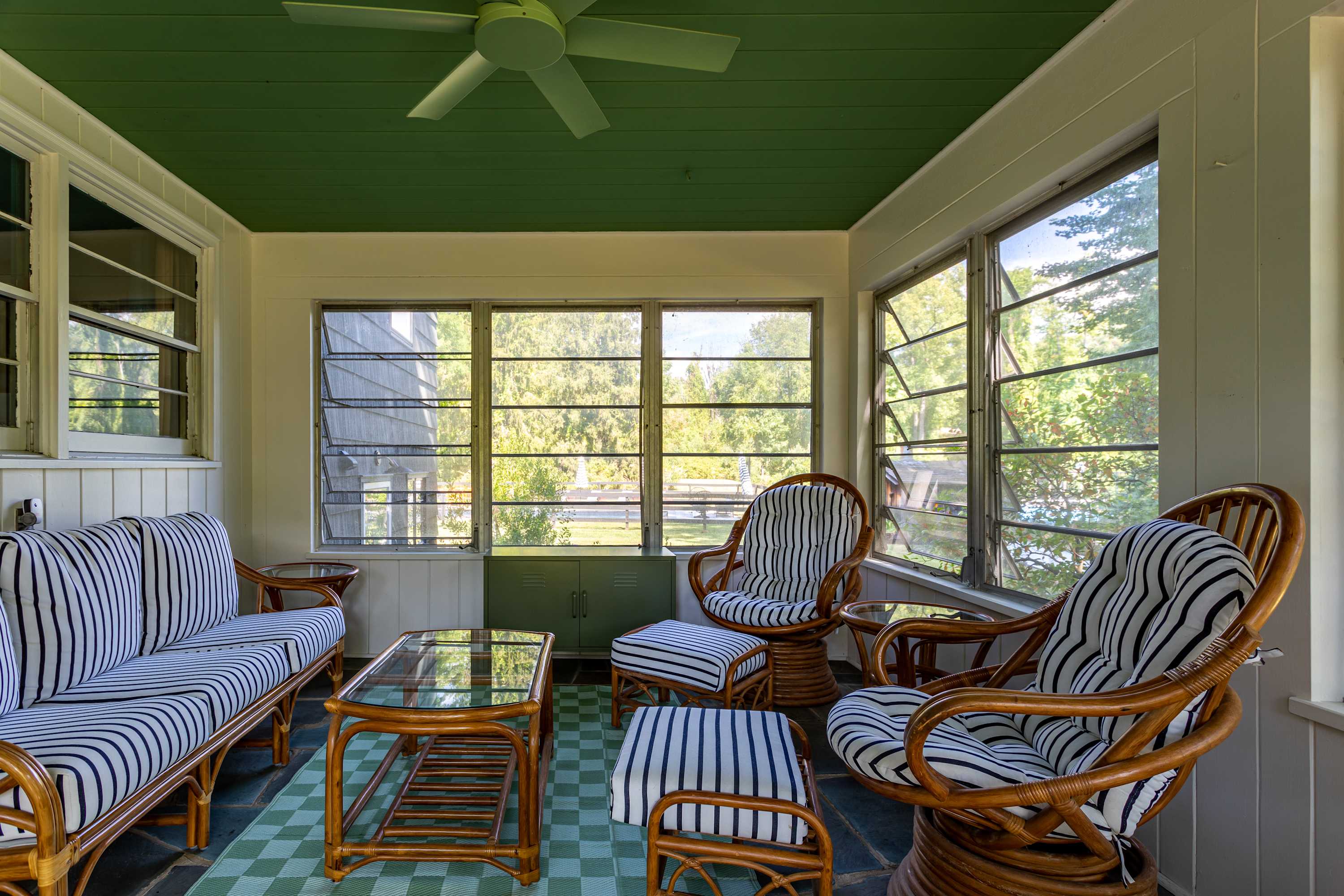 An enclosed porch with a vibrant green ceiling and checkered rug, furnished with rattan seating including a sofa, two armchairs, and an ottoman, all featuring black and white striped cushions, and a glass-top coffee table.
