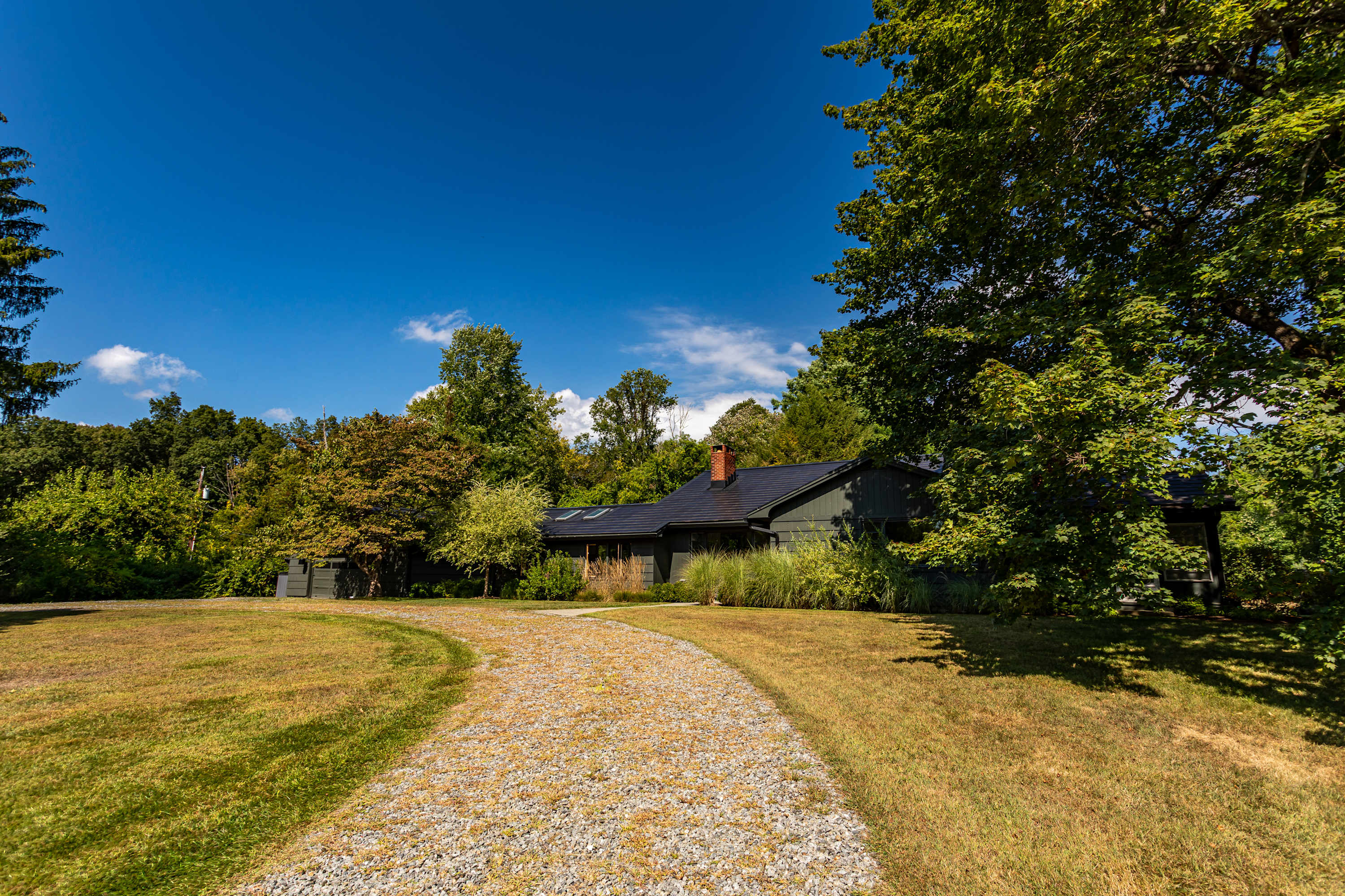 A gravel driveway curves through a grassy yard towards a dark-colored, single-story house with a dark roof and a brick chimney, surrounded by lush green trees under a clear blue sky.