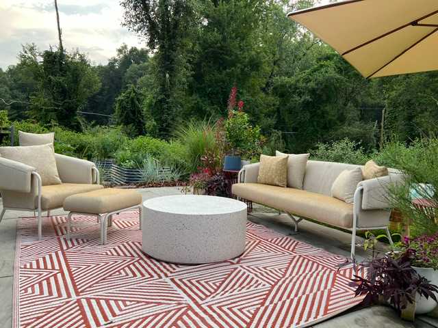 An inviting outdoor patio with a red and white geometric rug, a round concrete coffee table, and comfortable outdoor seating including a sofa and armchairs. Lush greenery and a large umbrella provide shade.