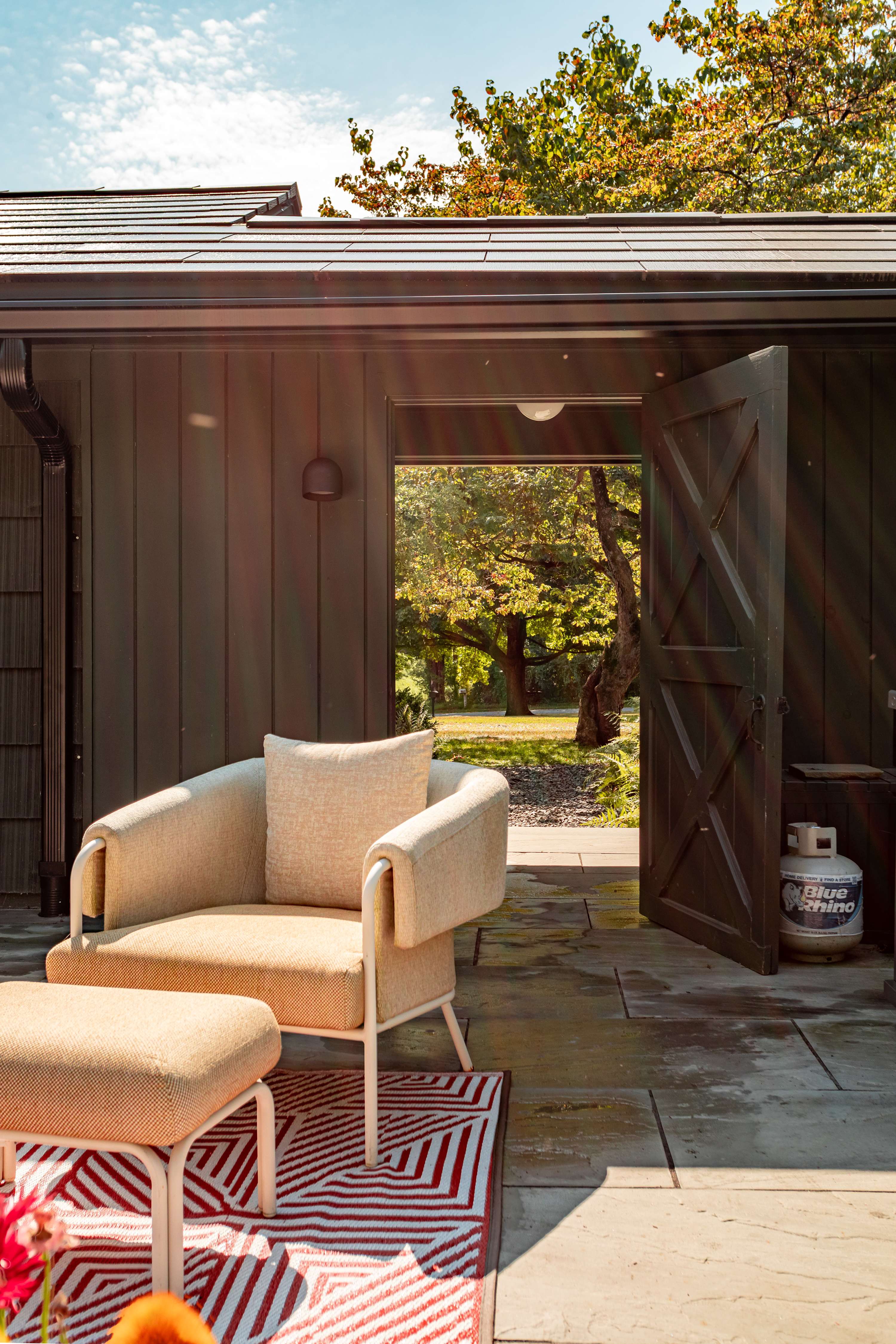 A cozy outdoor seating area on a stone patio, featuring a light beige armchair and ottoman on a red and white geometric rug, with a dark barn-style building and open door leading to a lush green landscape.