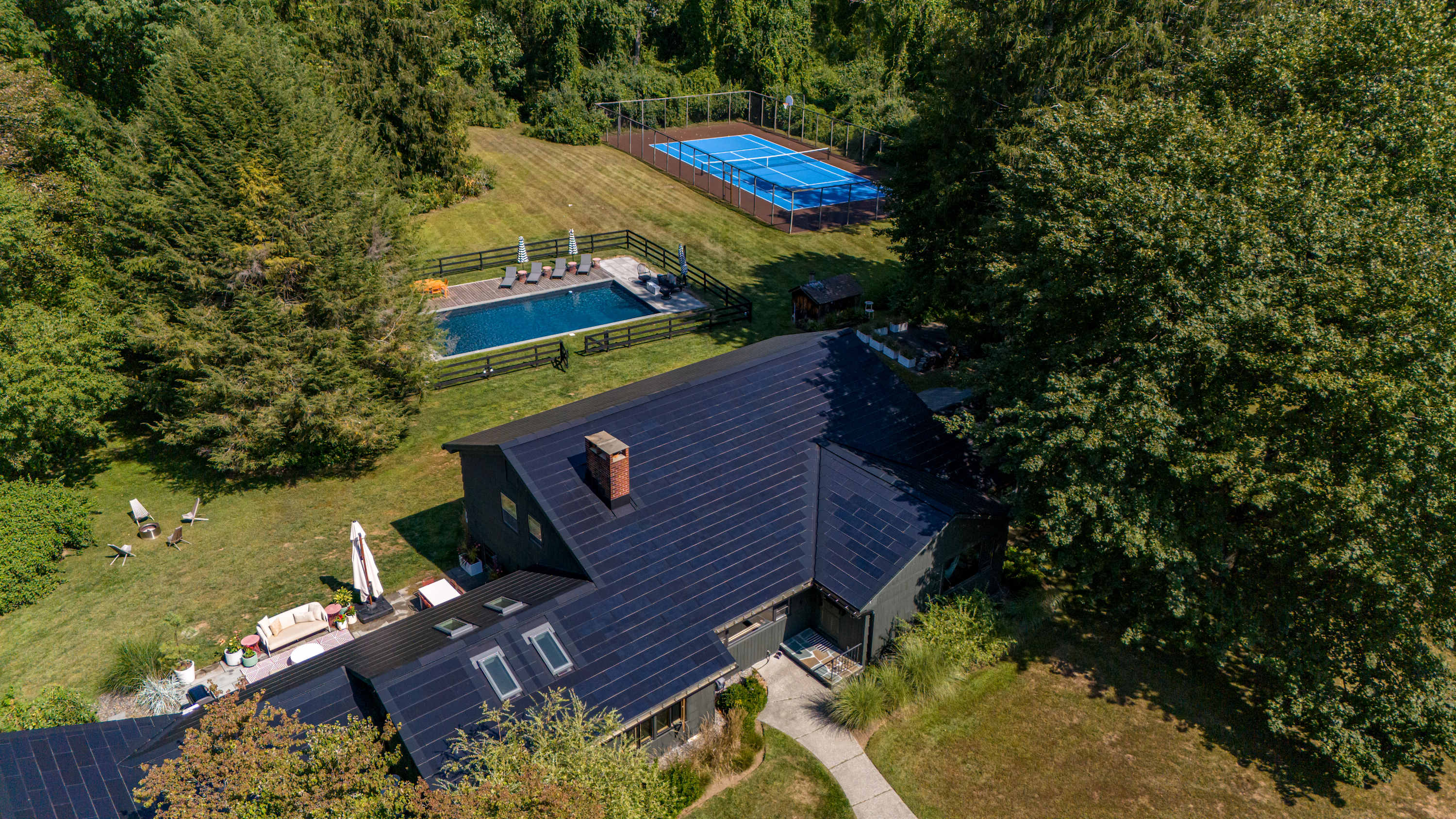 An aerial view showcases a dark-colored house with extensive solar panels, a fenced swimming pool area with lounge chairs, and a blue tennis court, all nestled within a lush green landscape of mature trees and expansive lawn.