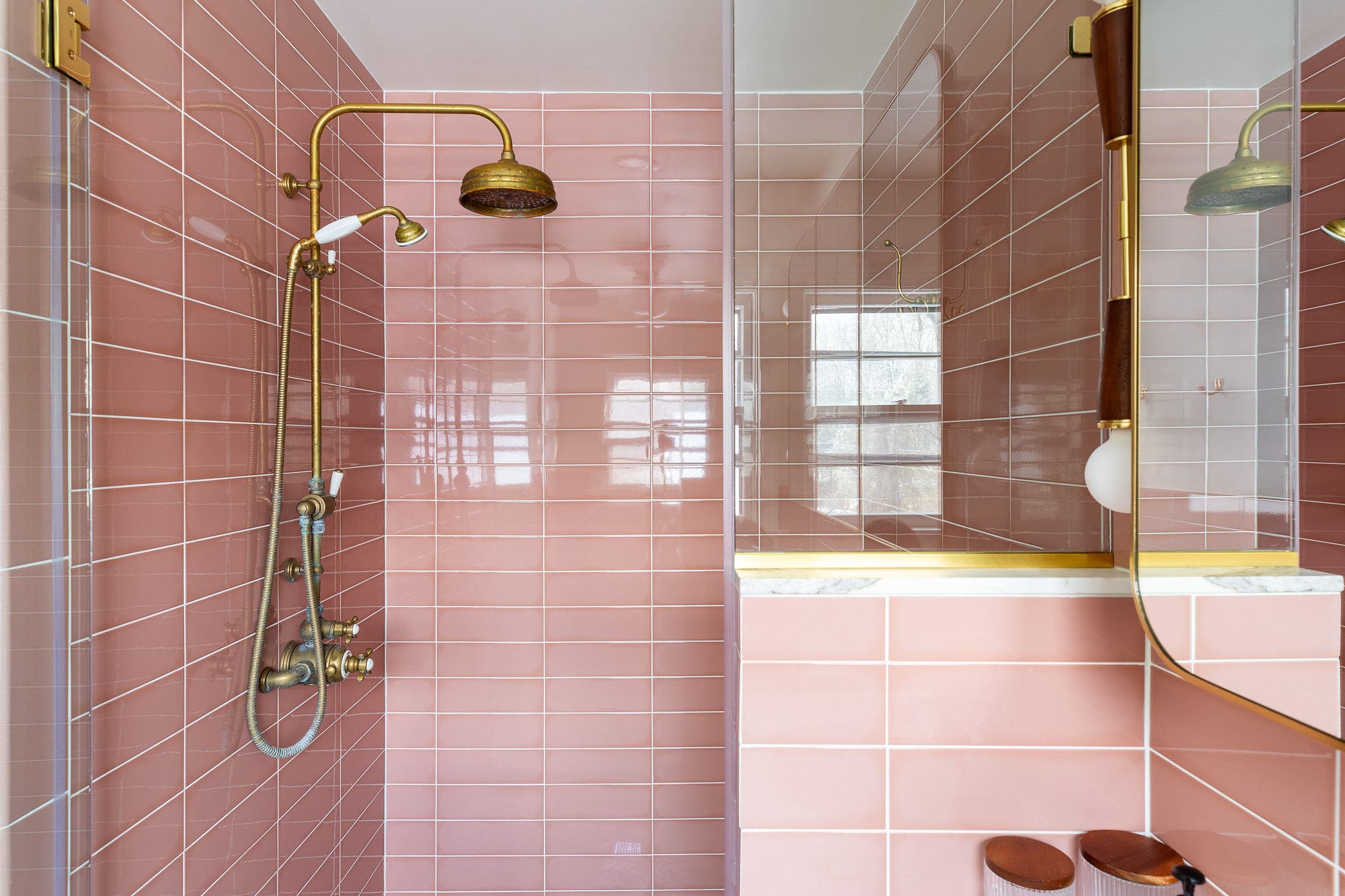 Close-up of the shower in the pink-tiled bathroom, featuring glossy pink subway tiles, a brass rain shower head and handheld sprayer, and a clear glass enclosure. A small marble ledge is also visible.