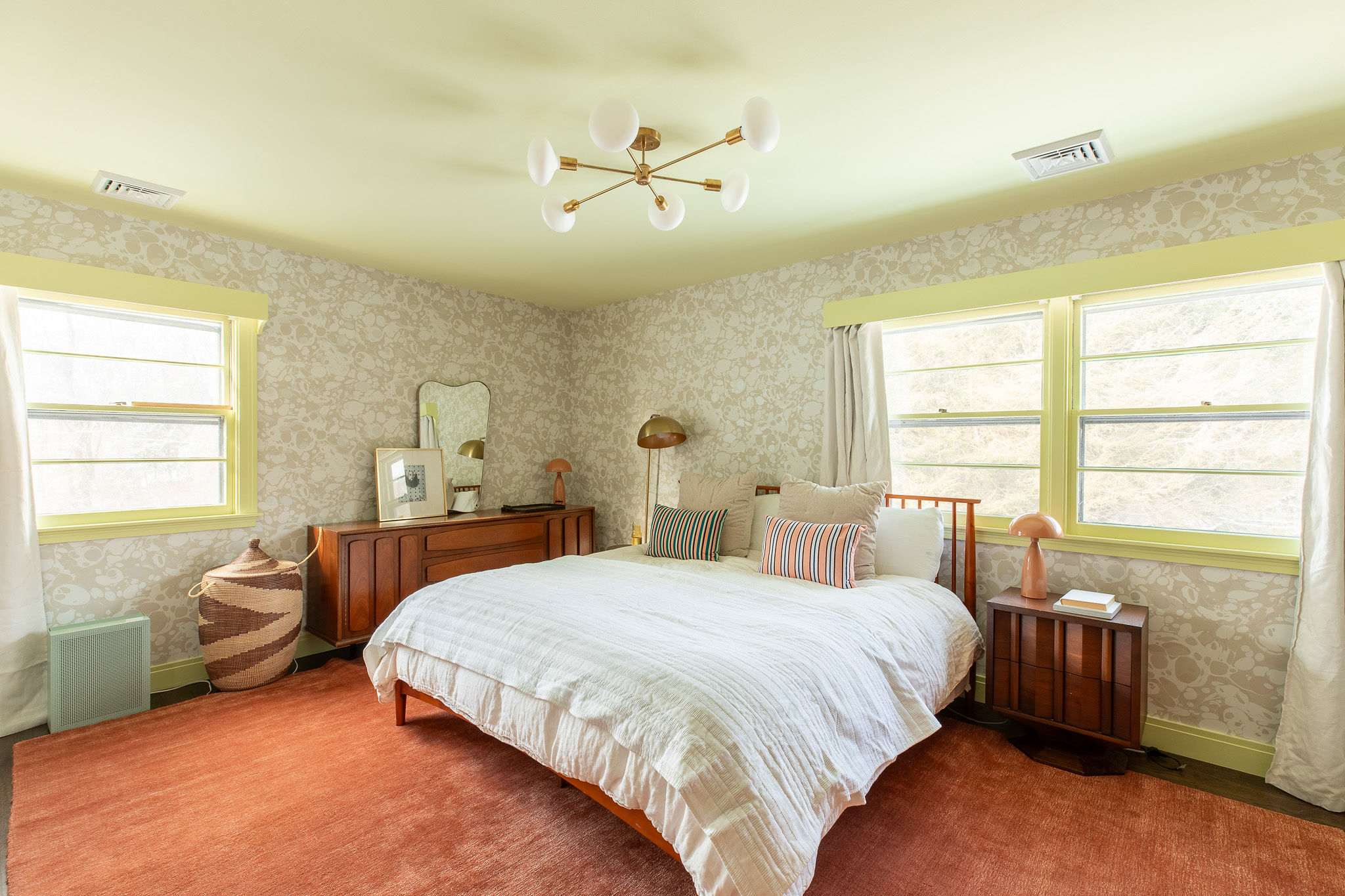 Bright bedroom with patterned wallpaper and light green trim, featuring a wooden bed with white bedding and striped pillows, a mid-century modern dresser, and a distinctive brass chandelier. An orange rug covers the floor.