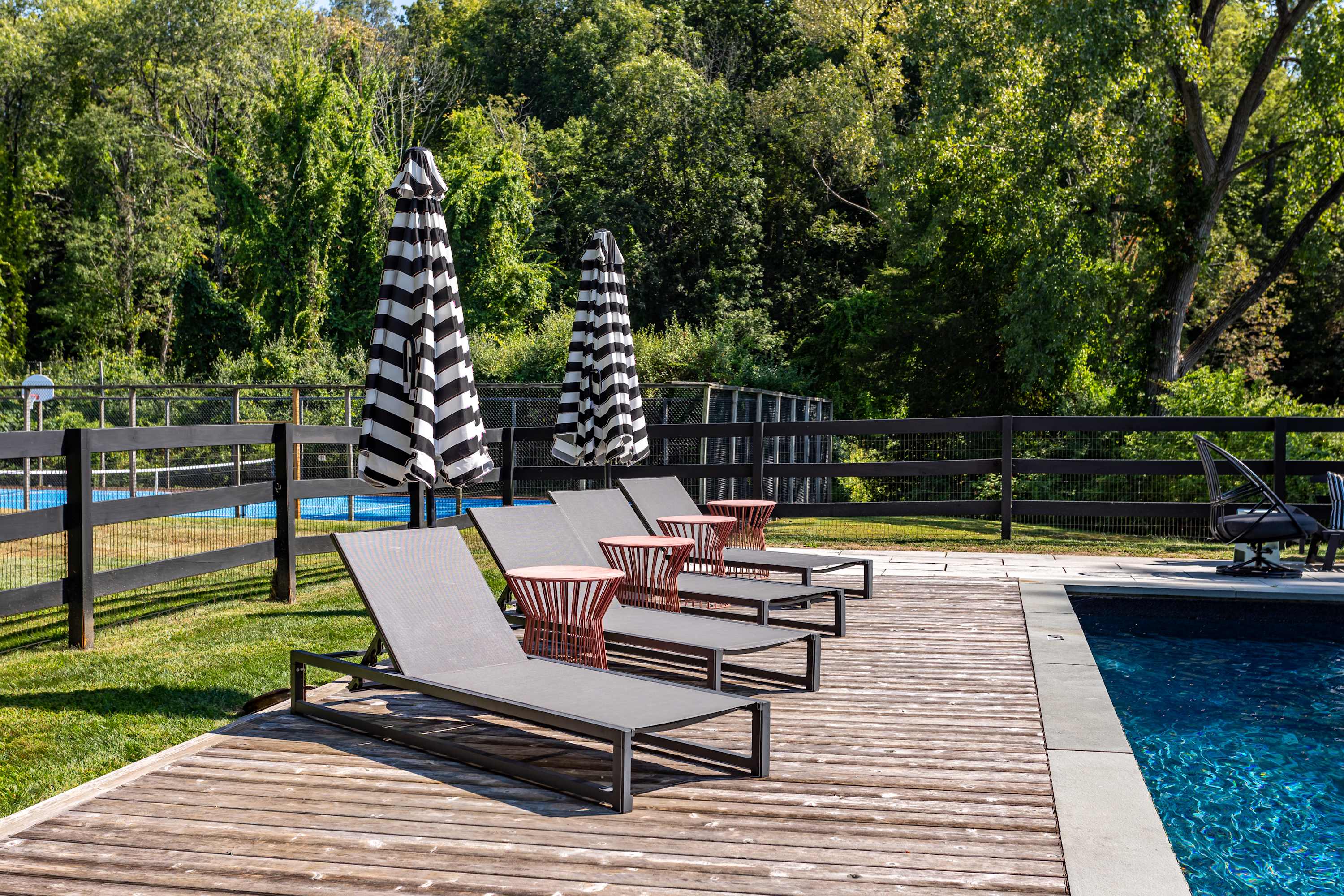 A row of four modern grey lounge chairs with small red side tables are neatly arranged on a wooden deck beside a dark blue swimming pool, shaded by two black and white striped umbrellas, with a black fence and lush trees in the background.