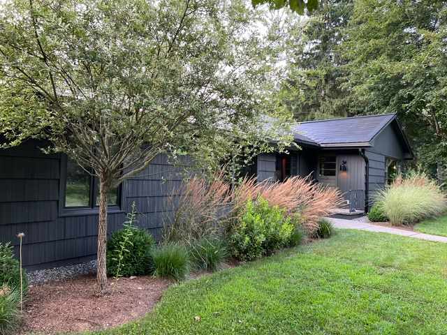 The front exterior of a dark gray house with shingle siding is partially obscured by a lush green lawn and various ornamental grasses and trees. A stone path leads to the entrance.
