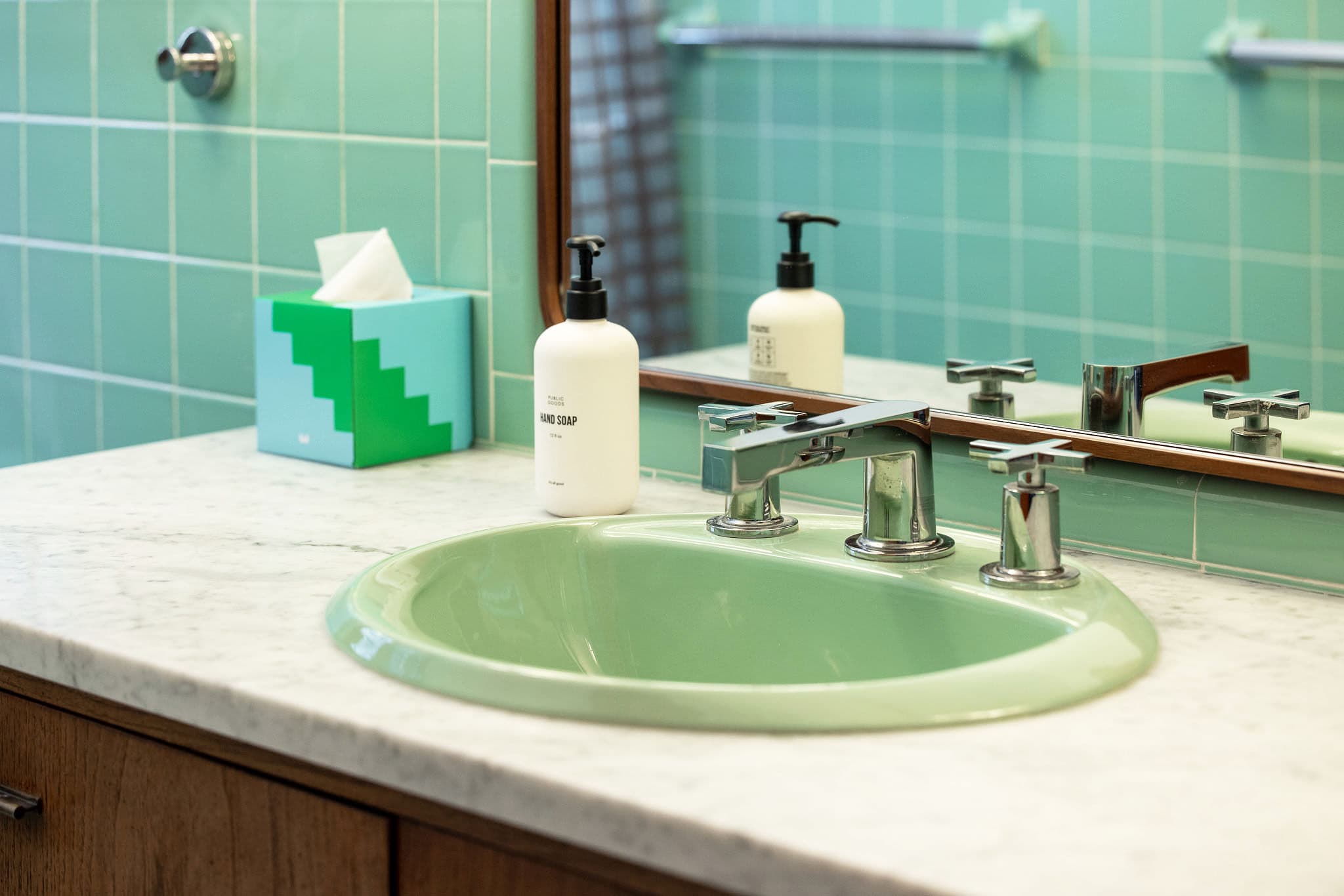 Close-up of the mint green sink set into a white marble countertop, featuring a chrome faucet with cross handles and a modern hand soap dispenser. A blue and green tissue box adds a pop of color.