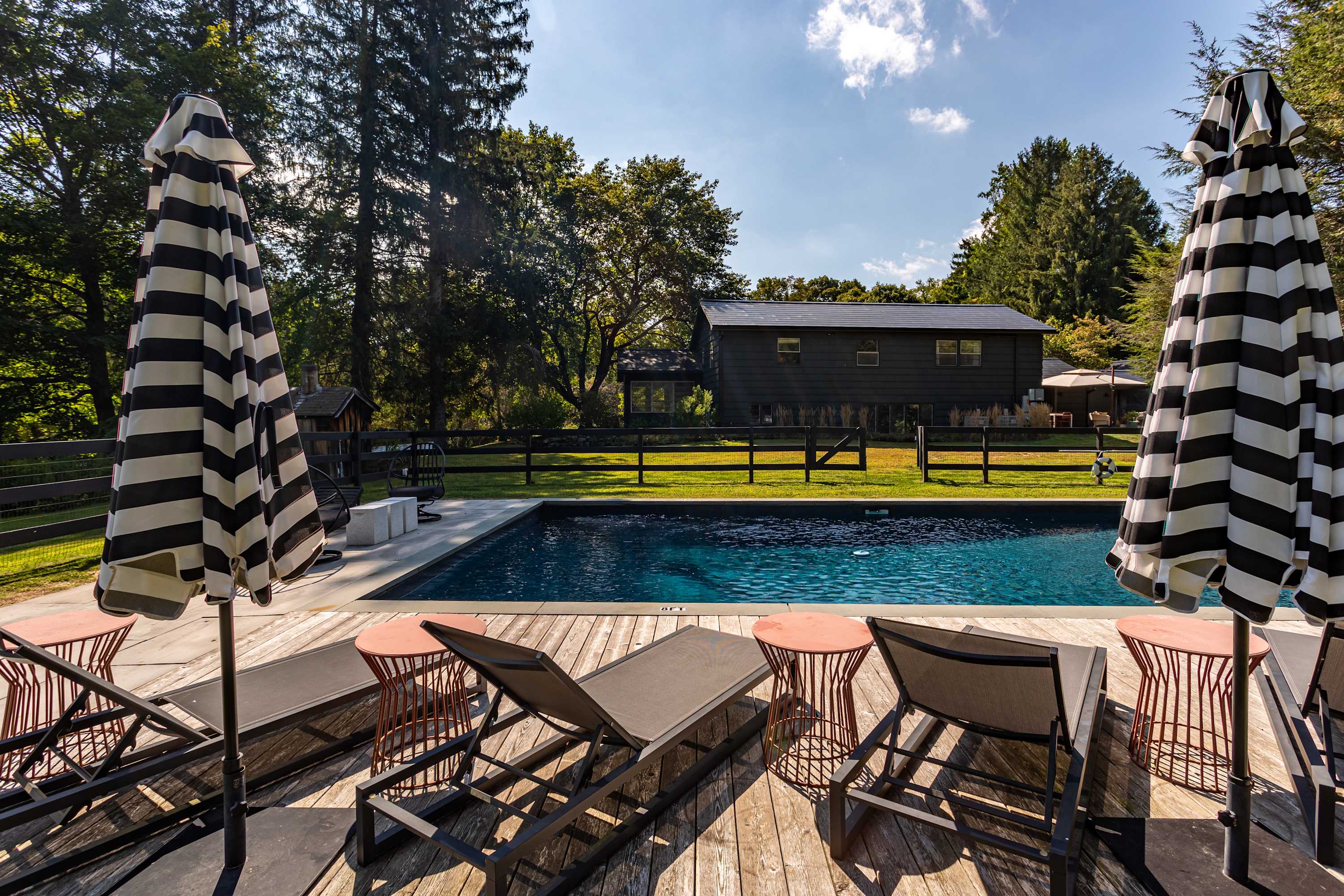 A view of the swimming pool area from a wooden deck, featuring several lounge chairs and two large black and white striped umbrellas, with a dark-sided house visible in the background beyond a black fence and lush trees.