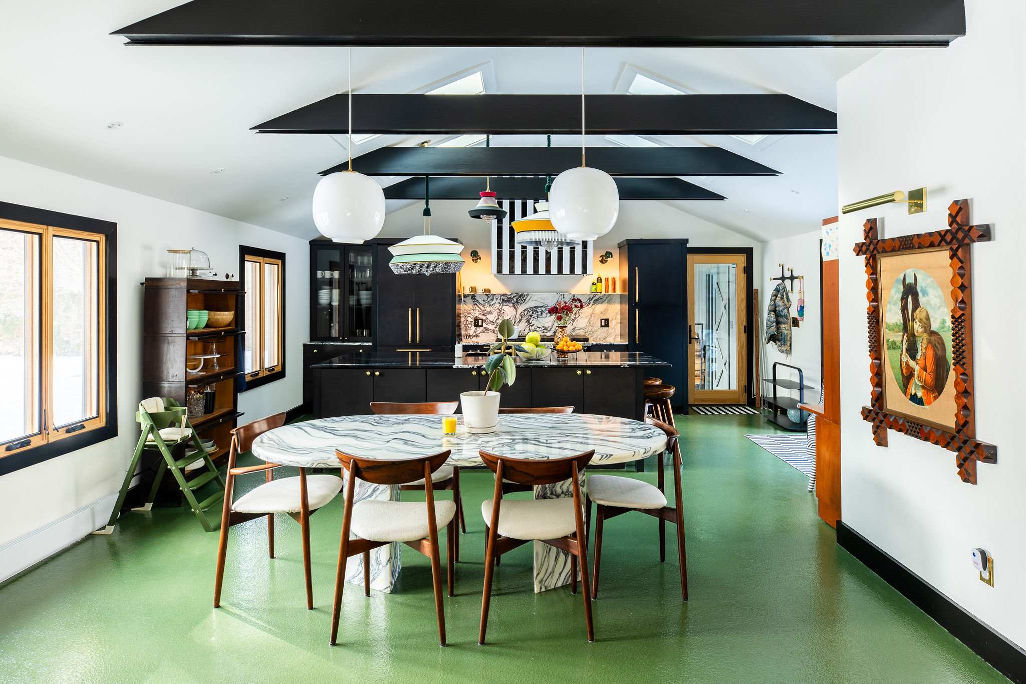 An open-concept dining and kitchen area features a large marble-topped dining table with wooden chairs, a black kitchen with a striped range hood, and a vibrant green epoxy floor.
