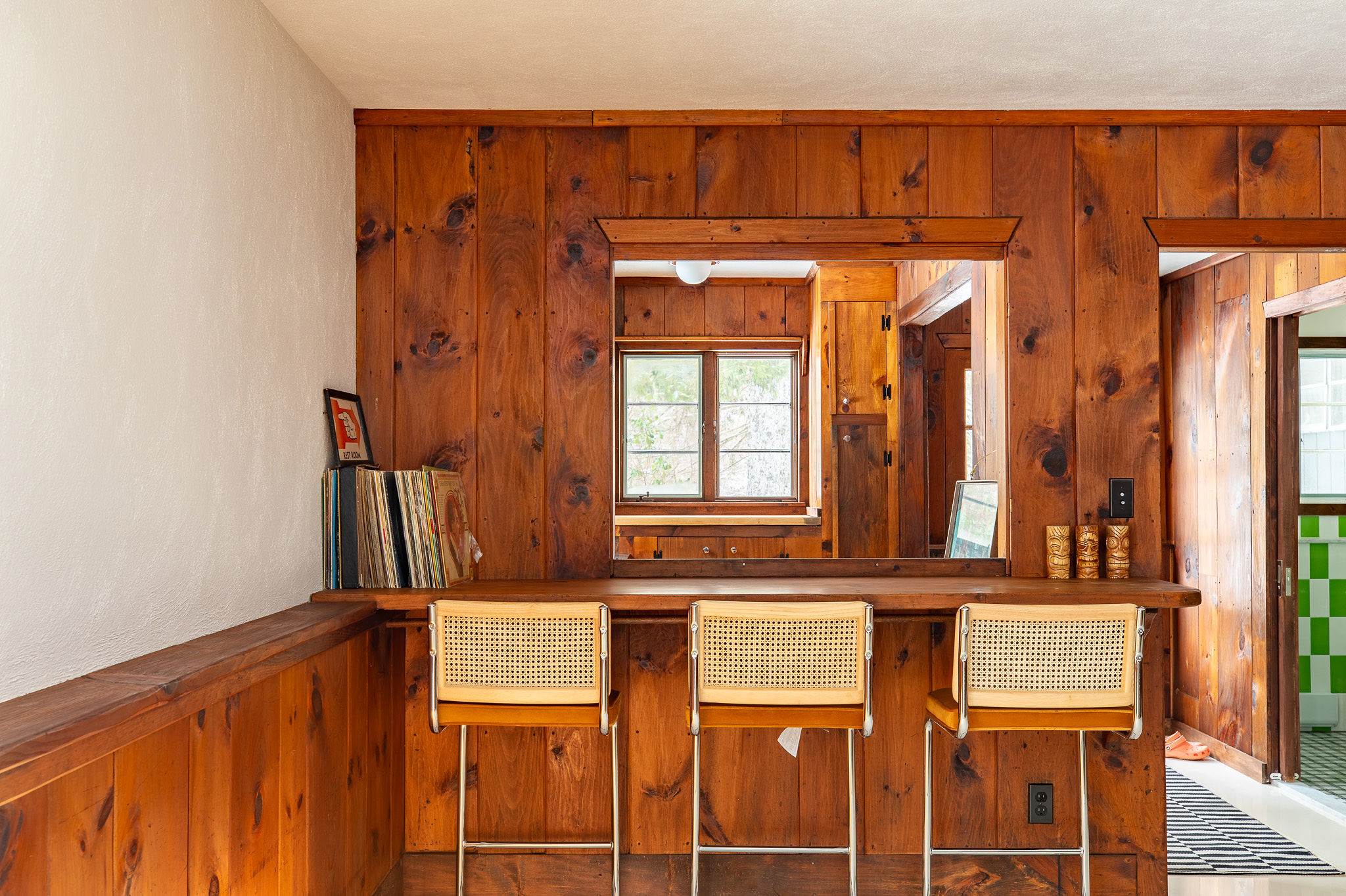 A rustic bar area integrated into the wood-paneled living room, featuring a wooden countertop with three stylish cane-back bar stools and a window overlooking the outdoors. Records are displayed on a shelf to the left.