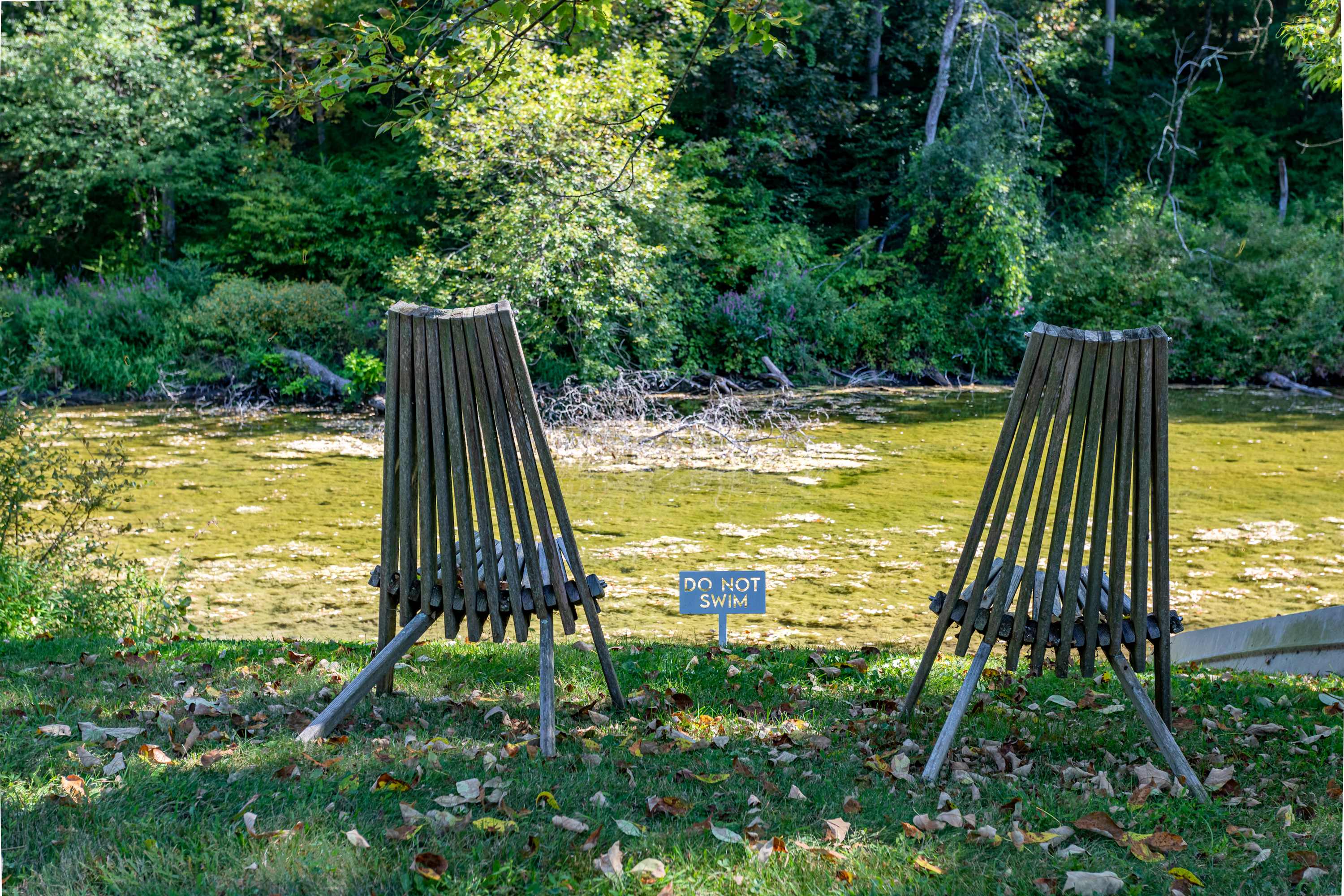 Two rustic wooden Adirondack chairs face a tranquil pond with green-tinged water, bordered by lush green trees and foliage, with a small 'DO NOT SWIM' sign placed between the chairs.