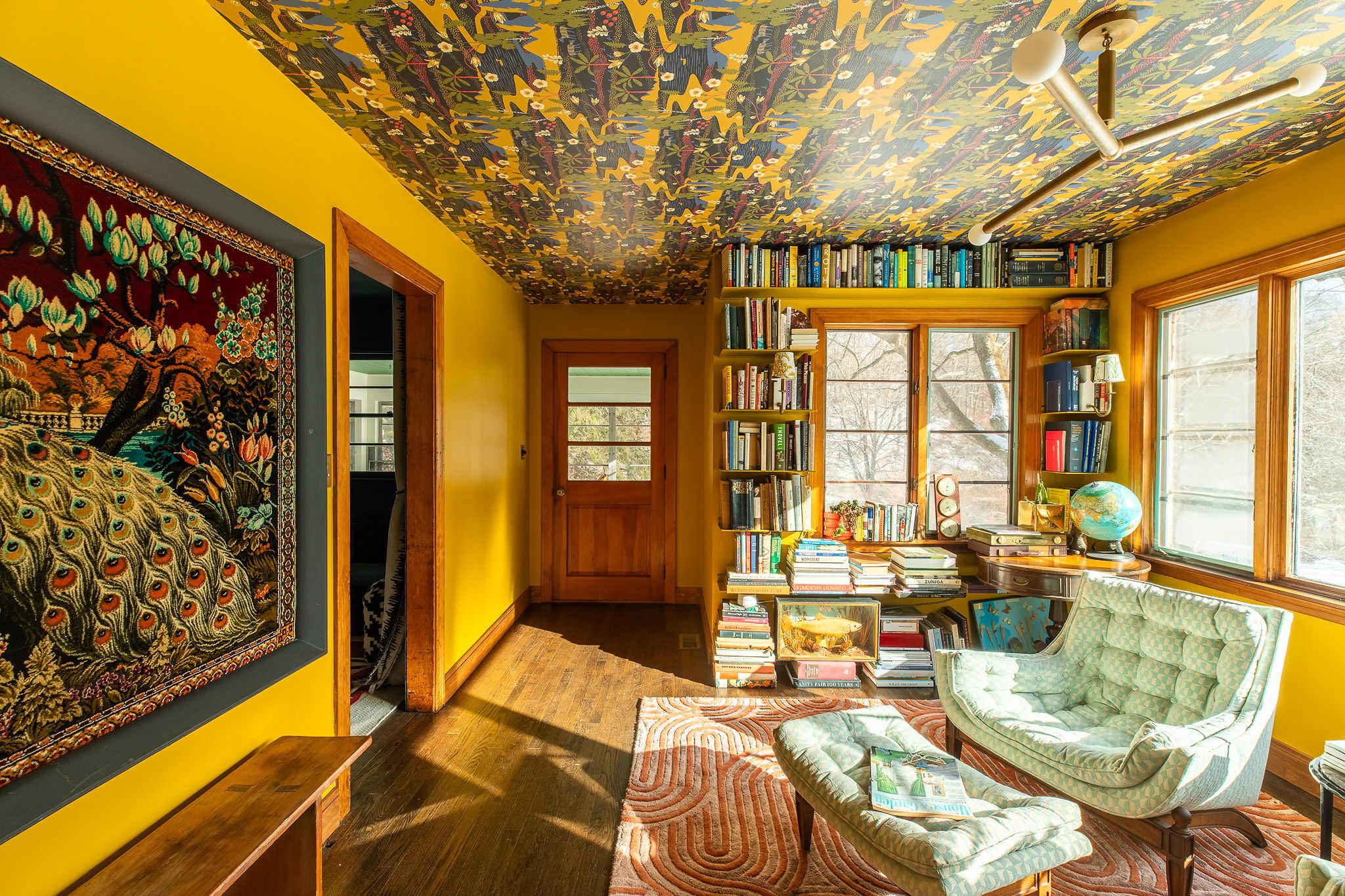 A cozy office or library room with bright yellow walls and a striking patterned wallpaper ceiling. Built-in bookshelves line a window, filled with books, while a light green tufted armchair and ottoman offer a comfortable reading nook. A large peacock tapestry hangs on one wall.