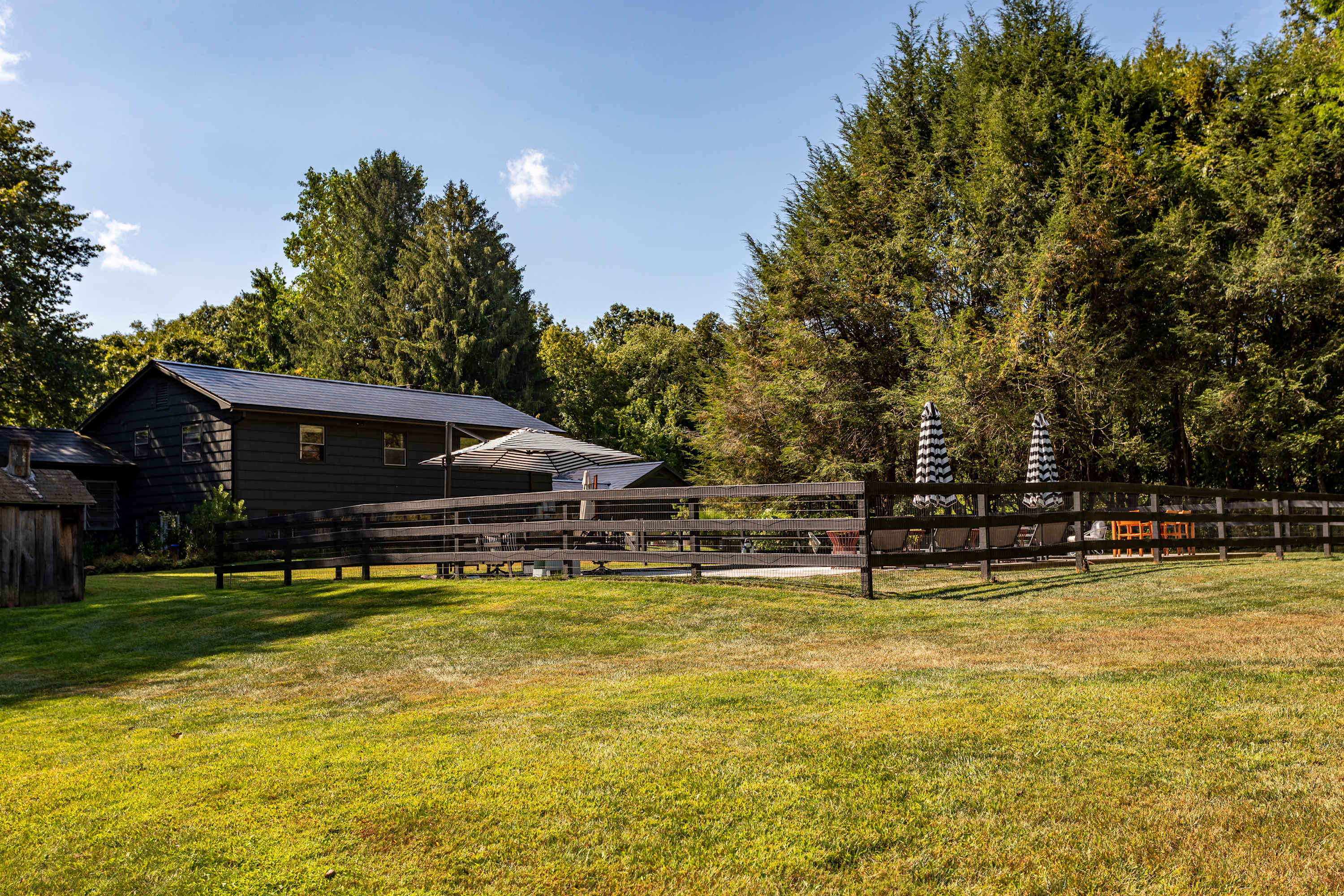 A dark-colored house with a patio area featuring outdoor furniture and umbrellas is visible behind a wooden fence, with a well-maintained green lawn in the foreground and tall evergreen trees in the background under a bright sky.