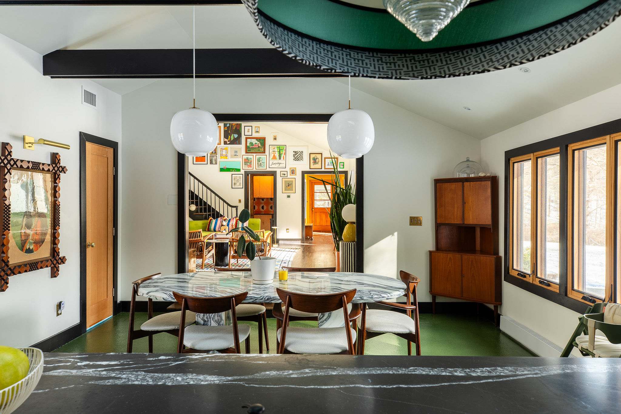 The dining area with its marble table and green floor opens into a bright living room, visible through a wide doorway, showcasing a gallery wall and a staircase.