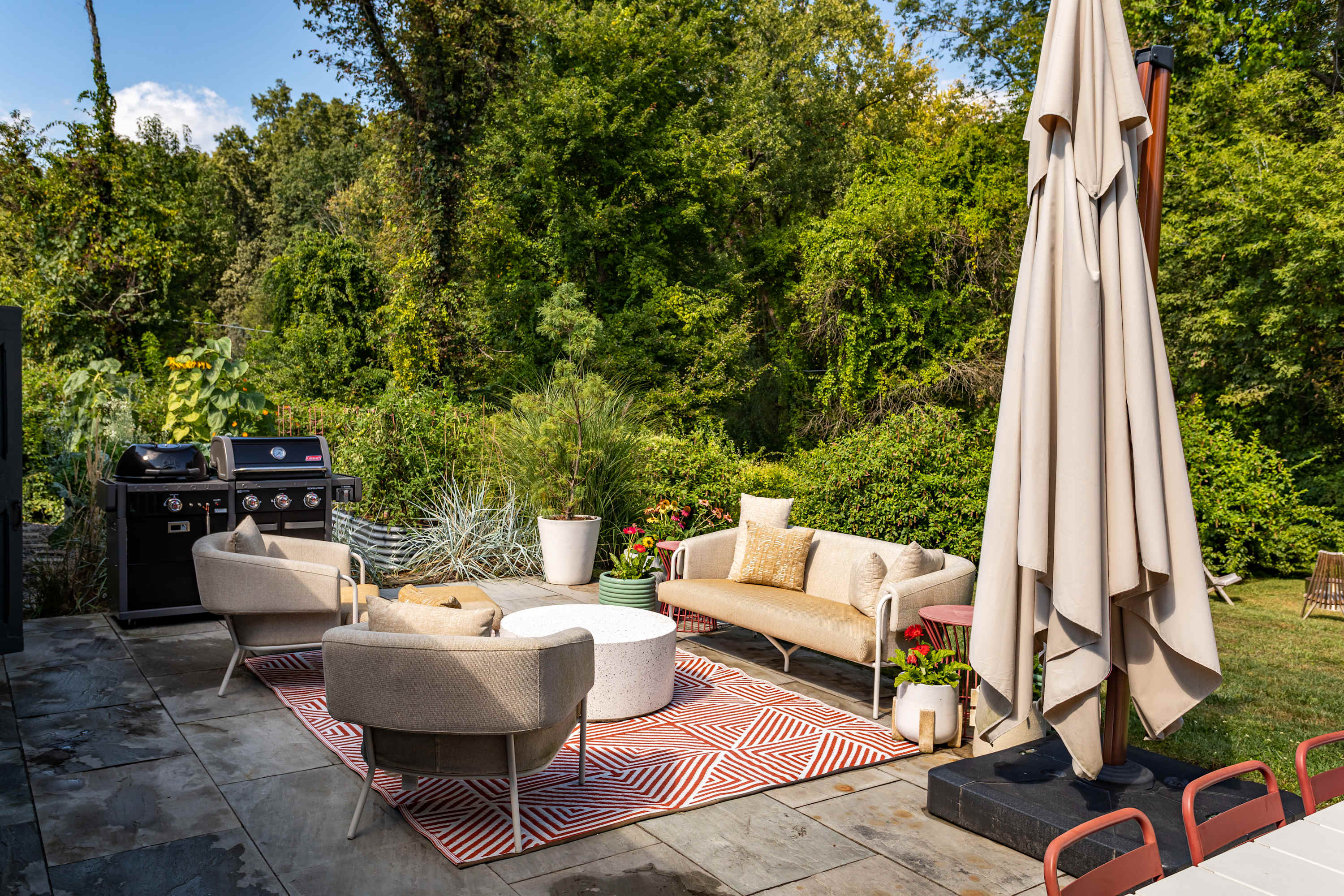 A spacious outdoor lounge area on a stone patio, featuring a beige sofa, two armchairs, and a round white coffee table on a red and white geometric rug, with a large BBQ grill and lush green foliage in the background.