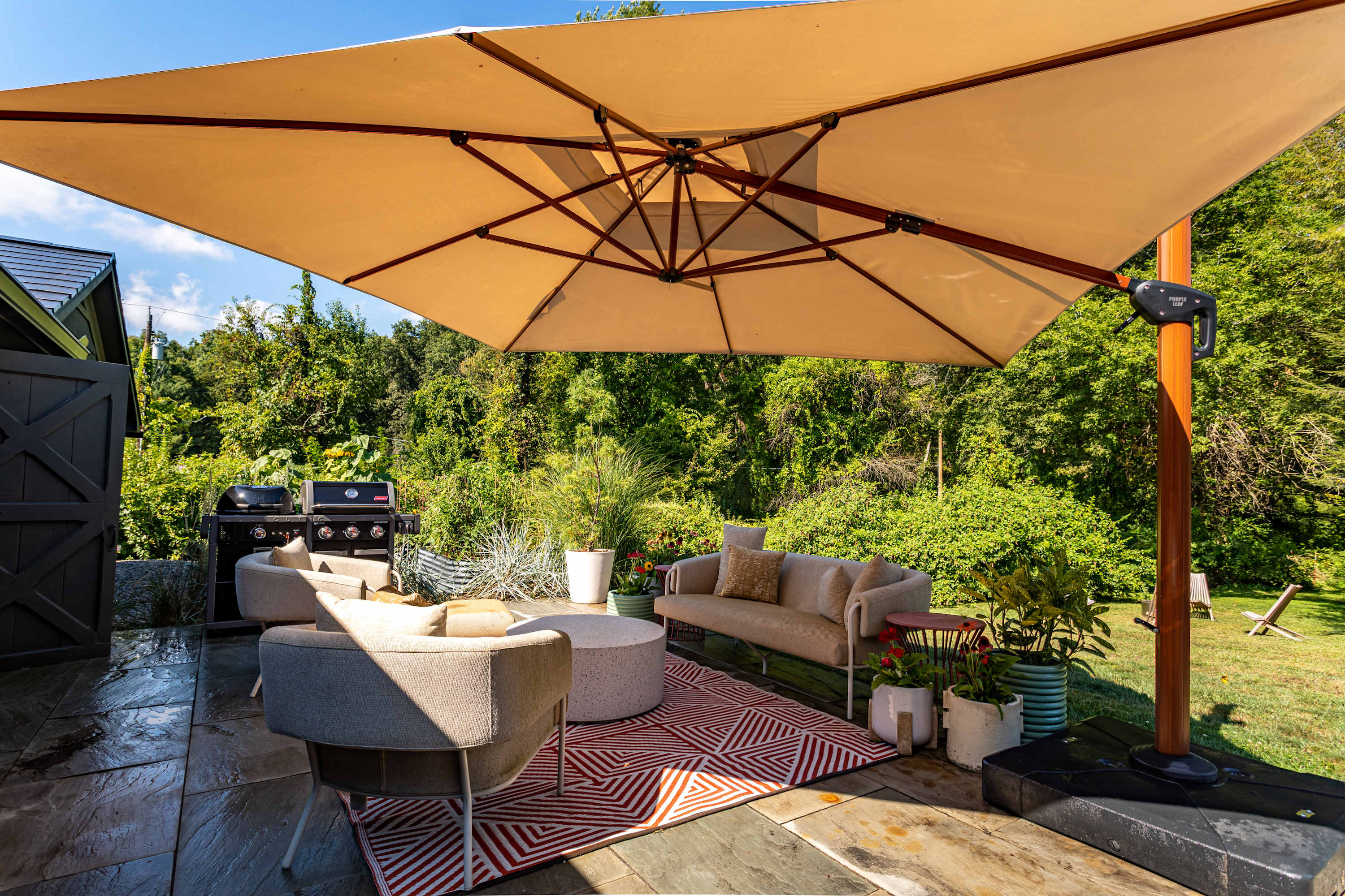 An inviting outdoor patio area features comfortable lounge seating and a large black BBQ grill, all shaded by a wide beige cantilever umbrella. Potted plants add greenery, and the area is surrounded by lush trees, suggesting a private setting.