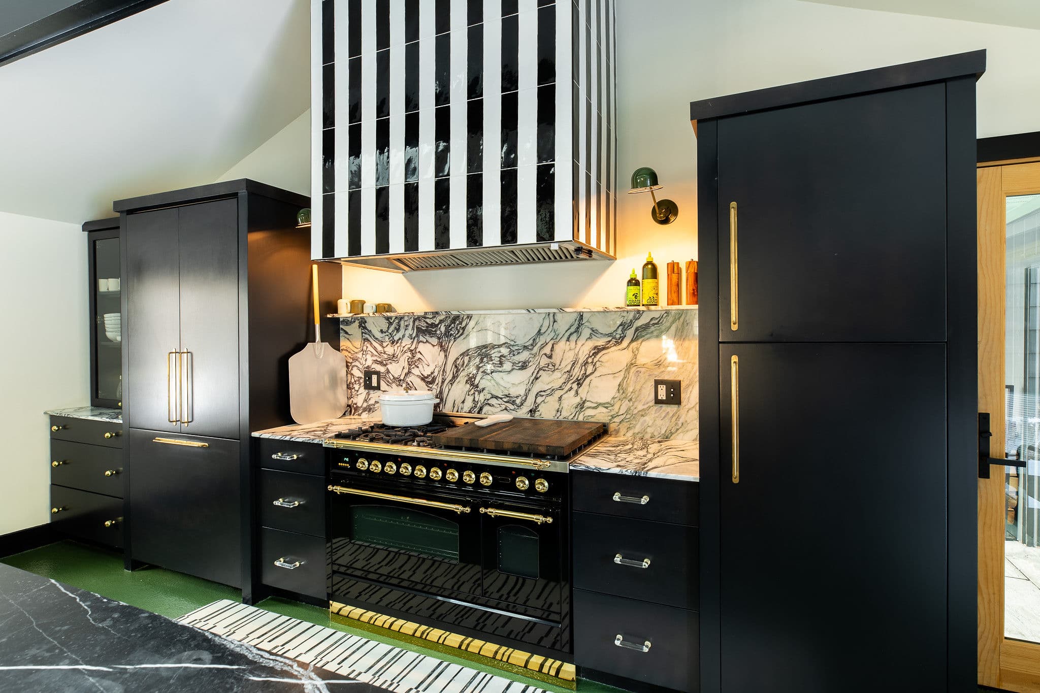 A stylish black kitchen features a large black and gold range, a striking black and white striped range hood, and a marble backsplash with intricate veining.