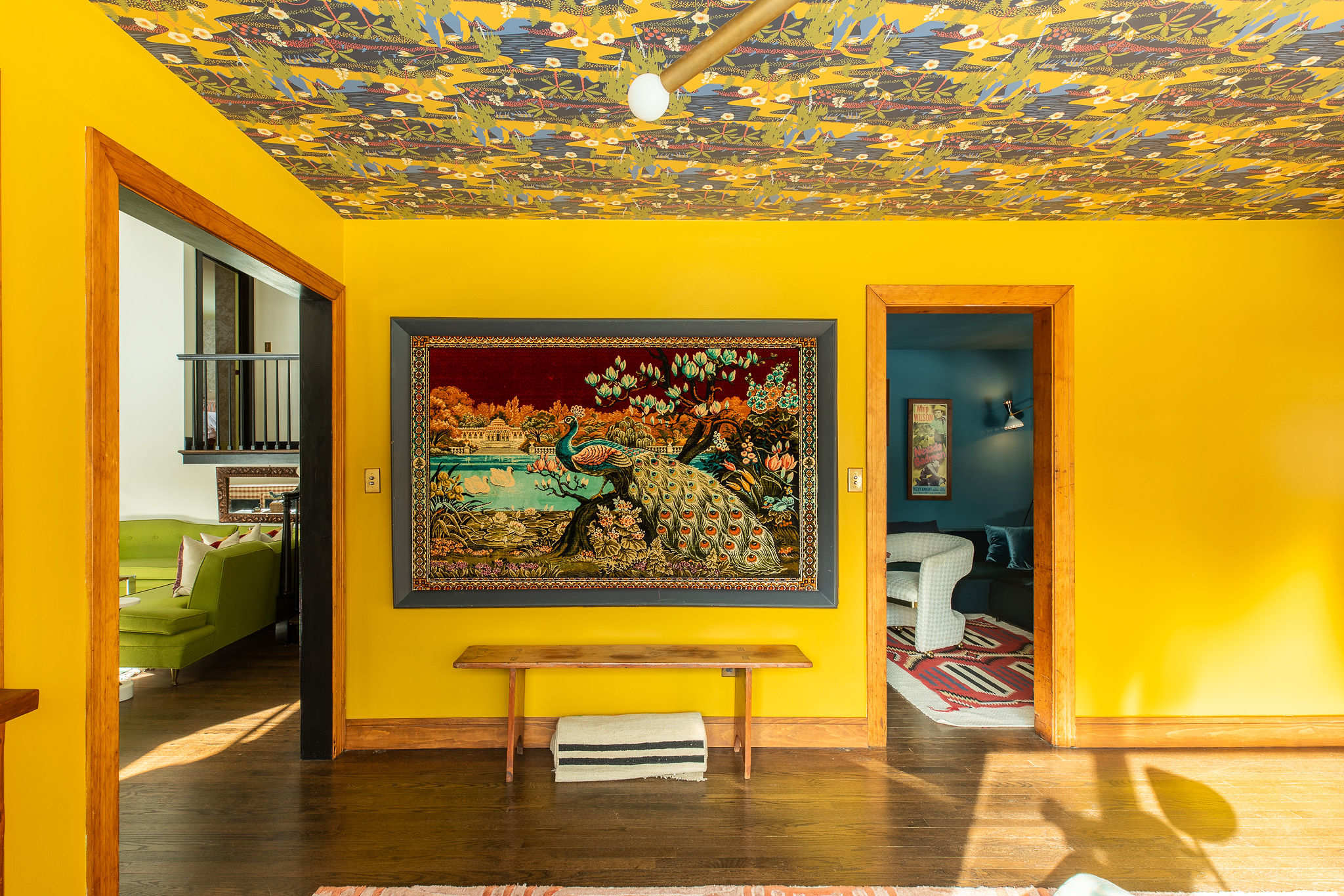 Bright yellow foyer with a bold patterned ceiling, featuring a large framed tapestry depicting peacocks and a landscape, positioned above a simple wooden bench. Doorways lead to other colorful rooms.