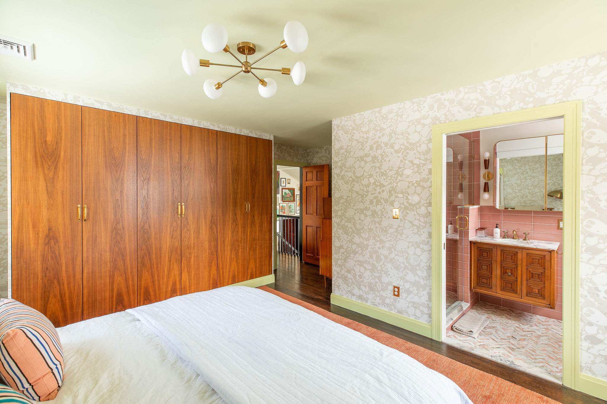 Bedroom view showcasing a wall of sleek wooden built-in wardrobes, a wooden bed with white bedding, and a doorway leading to a hallway. The room features patterned wallpaper and light green trim.