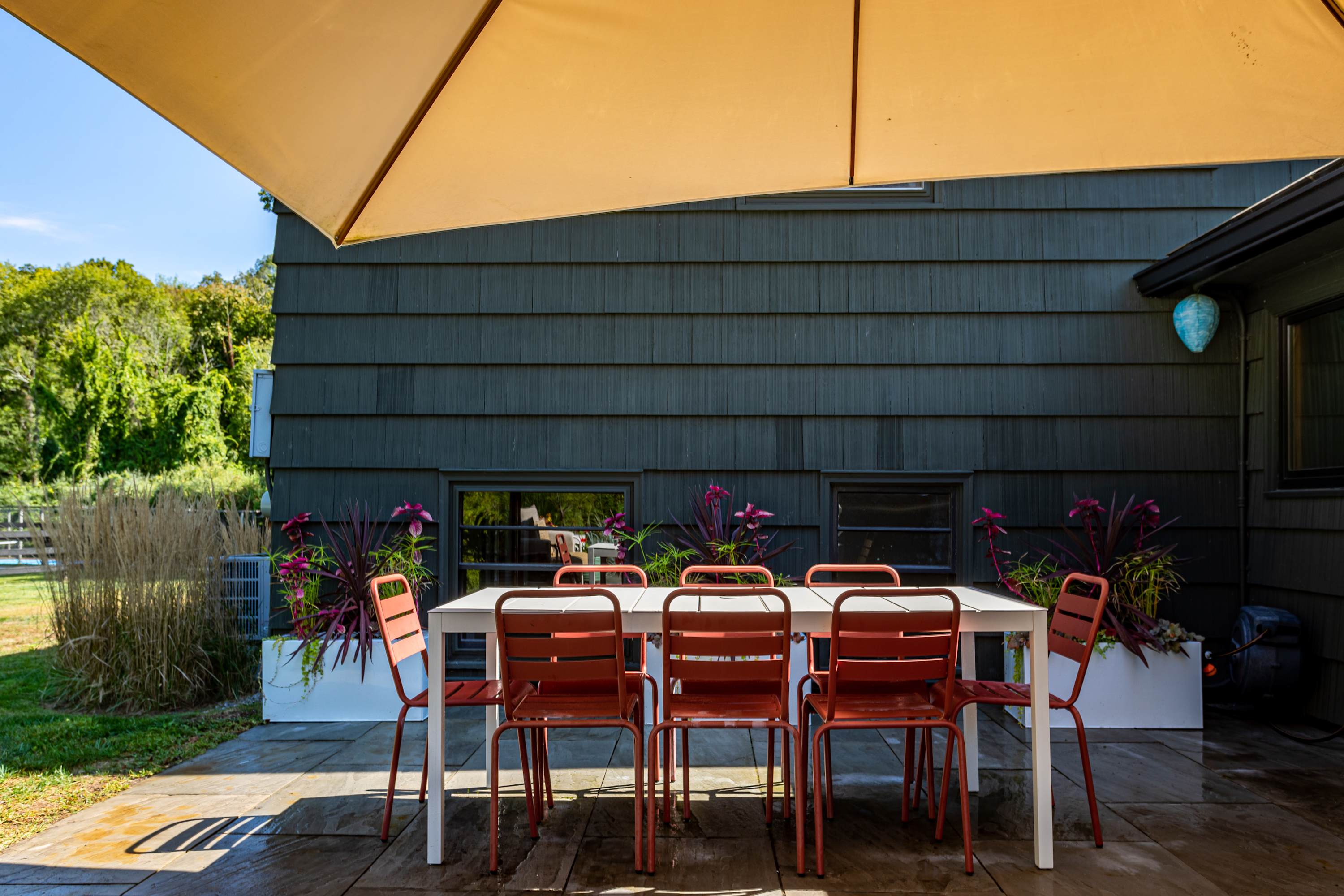 An outdoor dining setup on a stone patio, featuring a white rectangular table with red chairs, positioned under a large beige patio umbrella, next to a dark-sided building with windows and decorative planters.