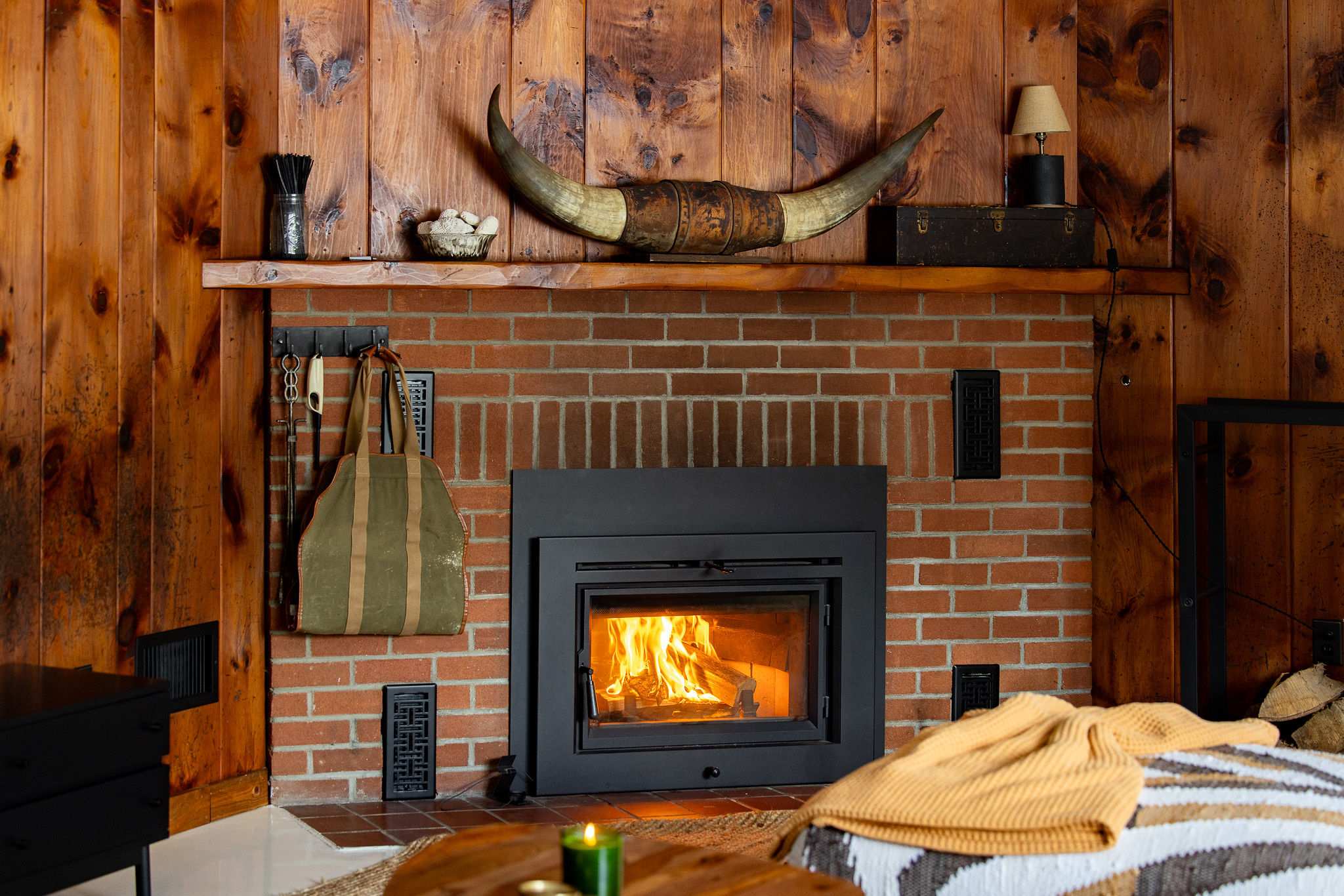 Close-up of the brick fireplace with a roaring wood-burning insert, set against warm wood-paneled walls. A rustic mantelpiece above features decorative bull horns and various accents.