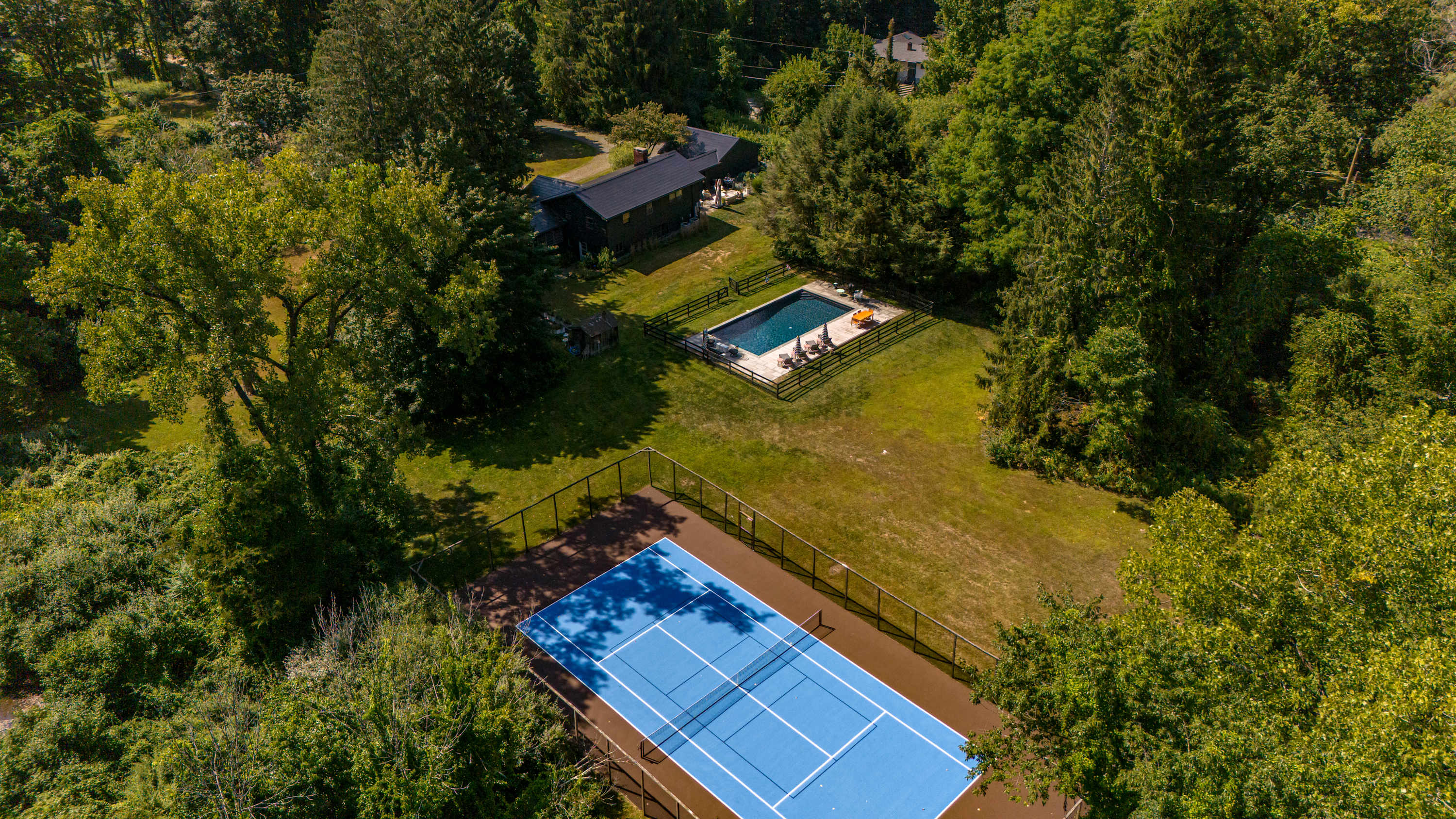 A high aerial view showcasing a sprawling property with a dark-roofed house, a fenced rectangular swimming pool, and a vibrant blue tennis court, all set within a vast expanse of green lawn and mature trees.
