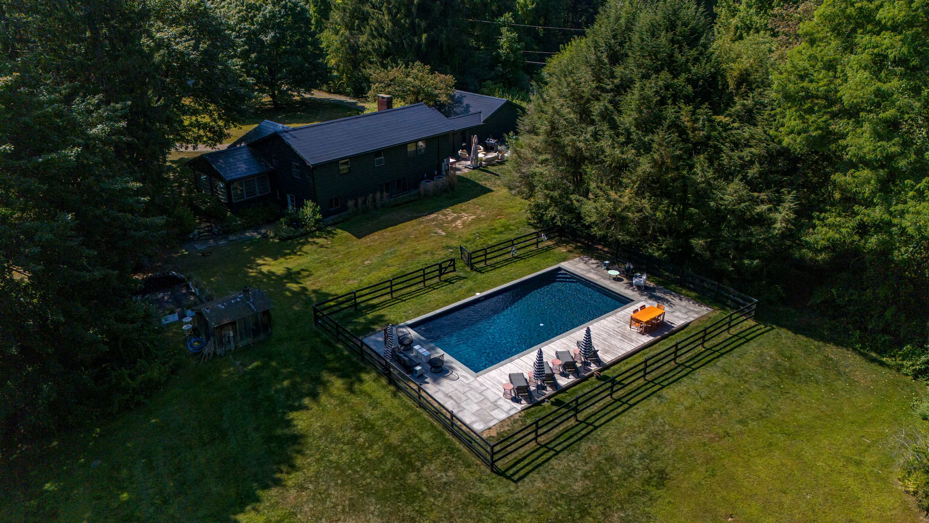 An aerial view of the property showing a dark-colored house, a fenced rectangular swimming pool with a surrounding deck, and a large grassy area, all nestled among abundant green trees.