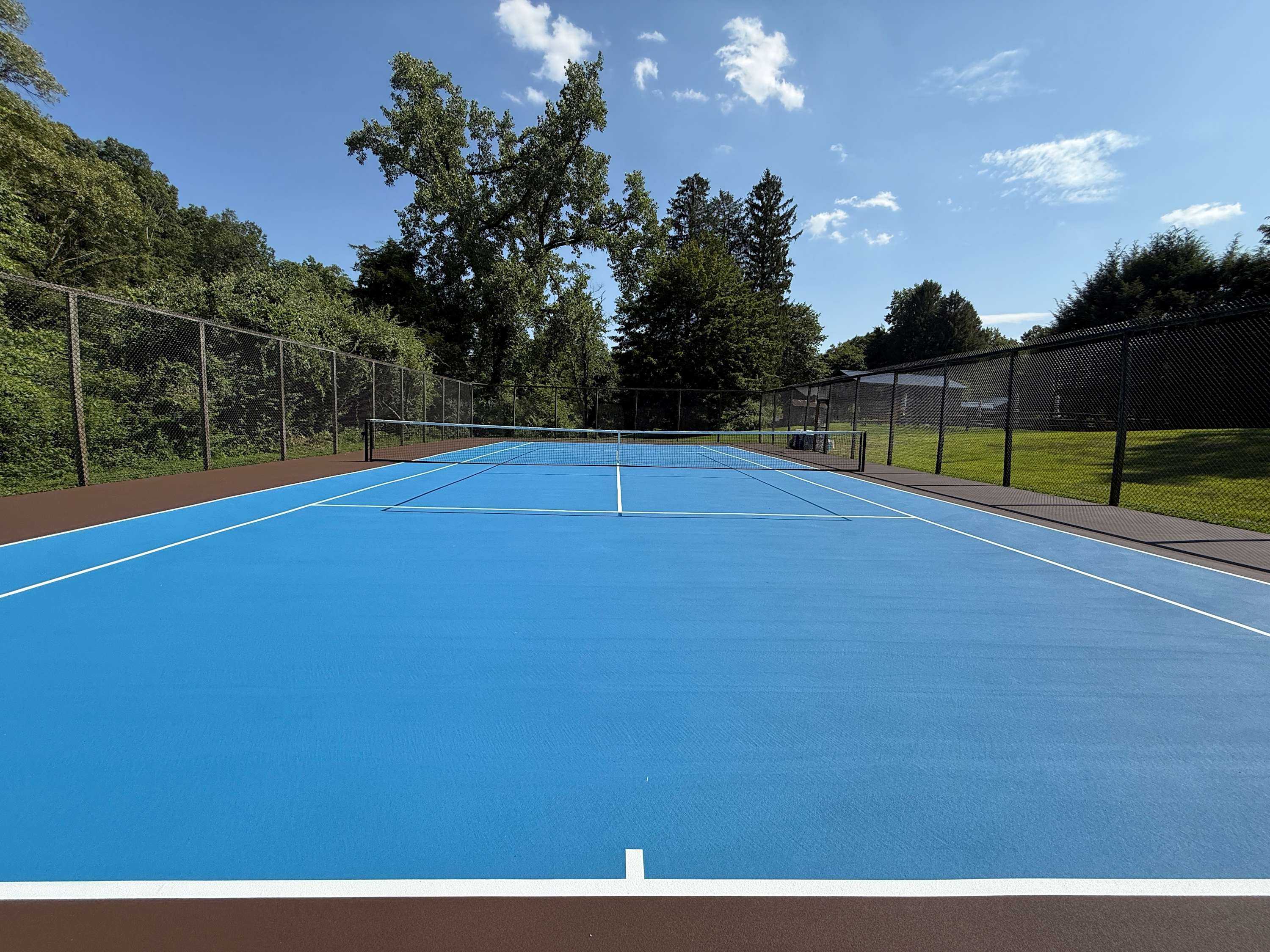 A wide-angle view of a pristine blue tennis court with white lines, enclosed by a black chain-link fence, set amidst lush green trees under a bright blue sky with scattered clouds.