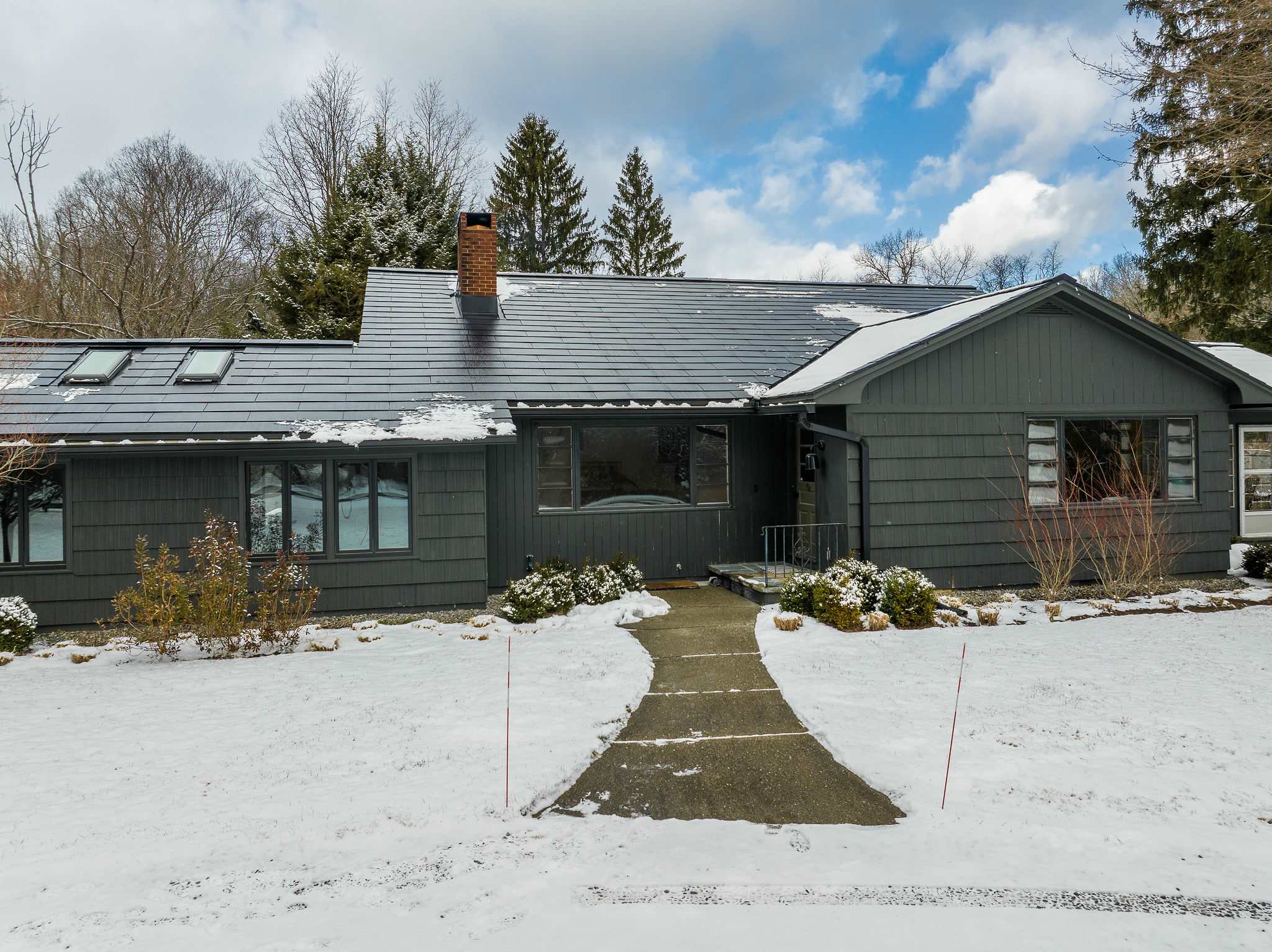 The front exterior of a dark gray house with a concrete walkway leading to the entrance, surrounded by snow-covered landscaping and evergreen bushes. The roof features skylights and a brick chimney, with tall trees in the background.