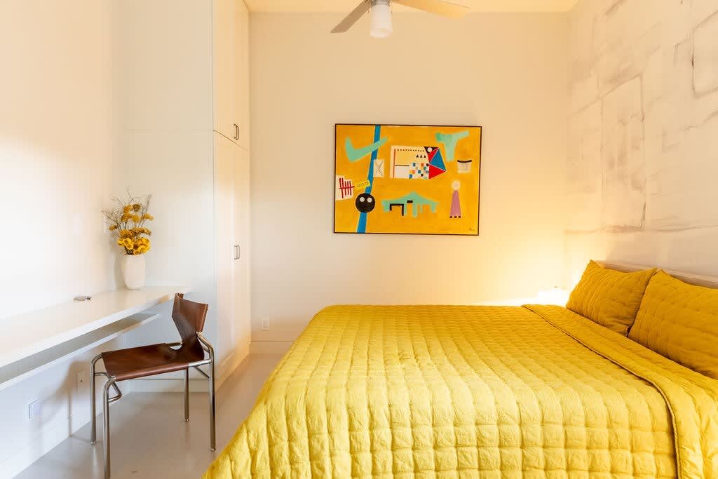A bright bedroom featuring a yellow quilted bed, a white built-in desk with a chair, a large abstract painting with yellow tones, and white built-in wardrobes, all under a ceiling fan.