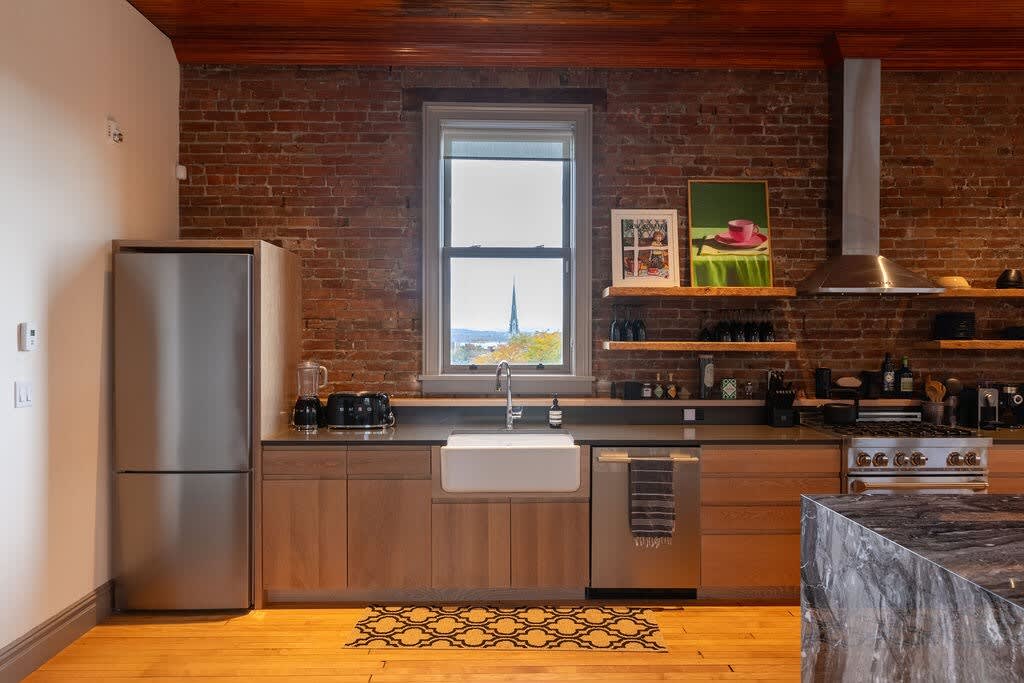 Detailed view of the kitchen counter with a white farmhouse sink, stainless steel appliances, light wood cabinetry, and exposed brick walls, with a window offering a distant view.