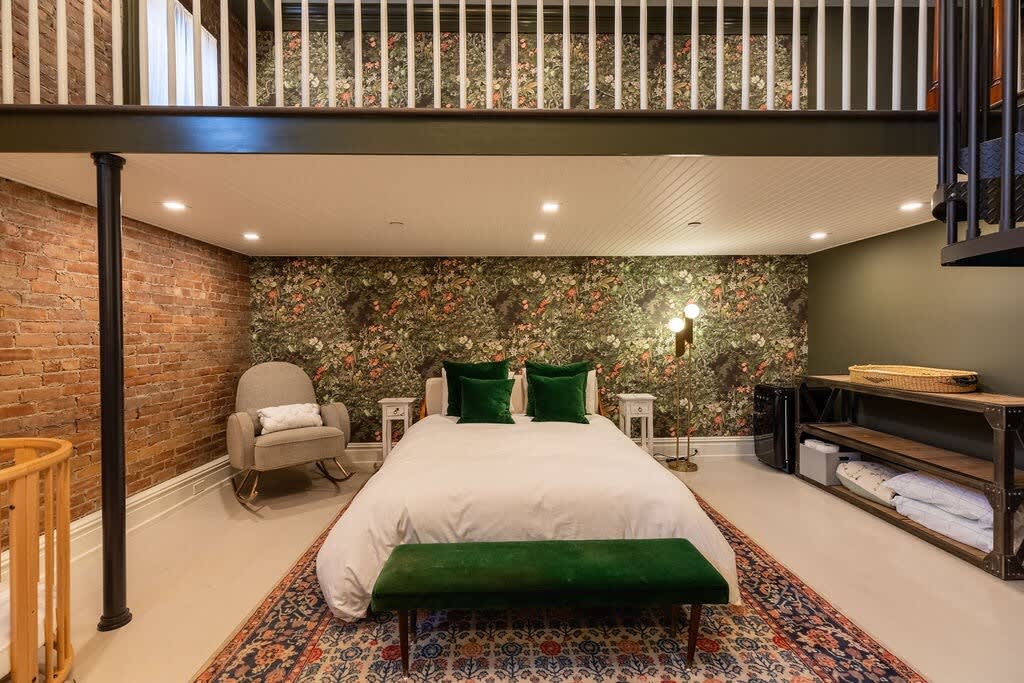 Expansive bedroom with a queen bed, green velvet bench, and a dark floral accent wall, complemented by an exposed brick wall. A wooden crib and a gray rocking chair are present, with a loft area visible above.