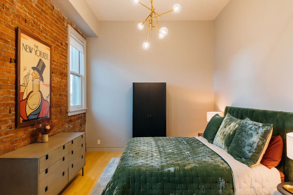 A modern bedroom with an exposed brick accent wall, a large window, a green velvet upholstered bed with matching bedding, a dark wardrobe, and a contemporary sputnik chandelier.