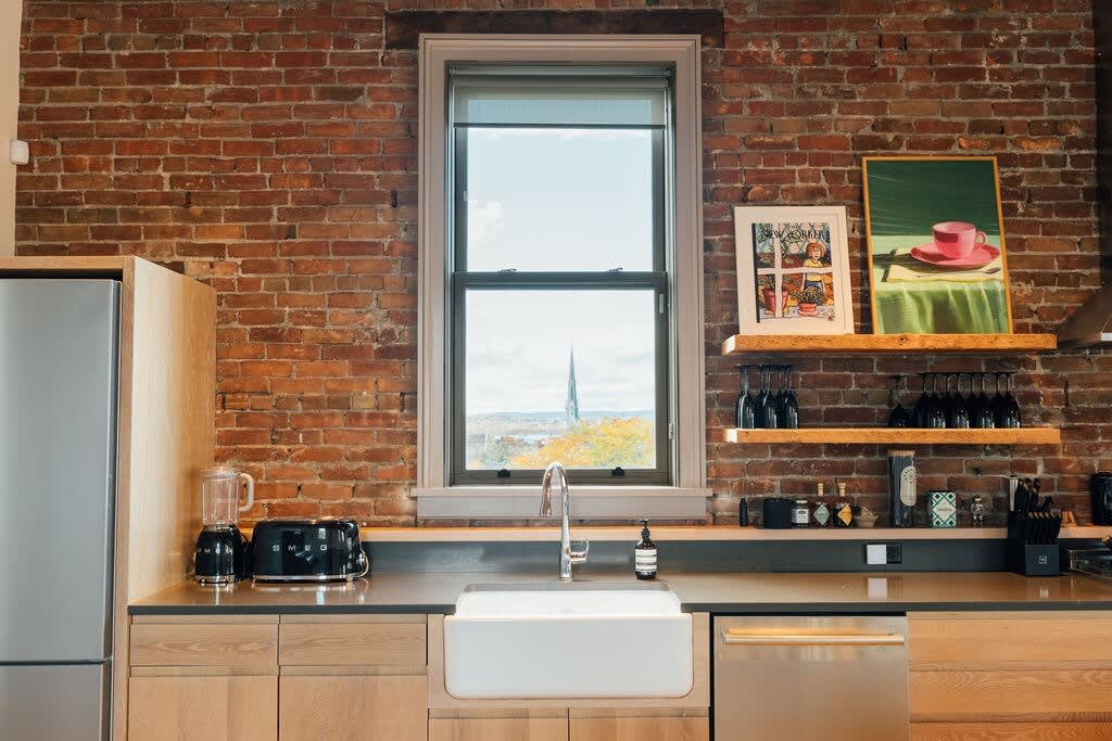 Bright kitchen counter with a white farmhouse sink, black small appliances, and open shelving against an exposed brick wall, featuring a window with a view of a distant spire.