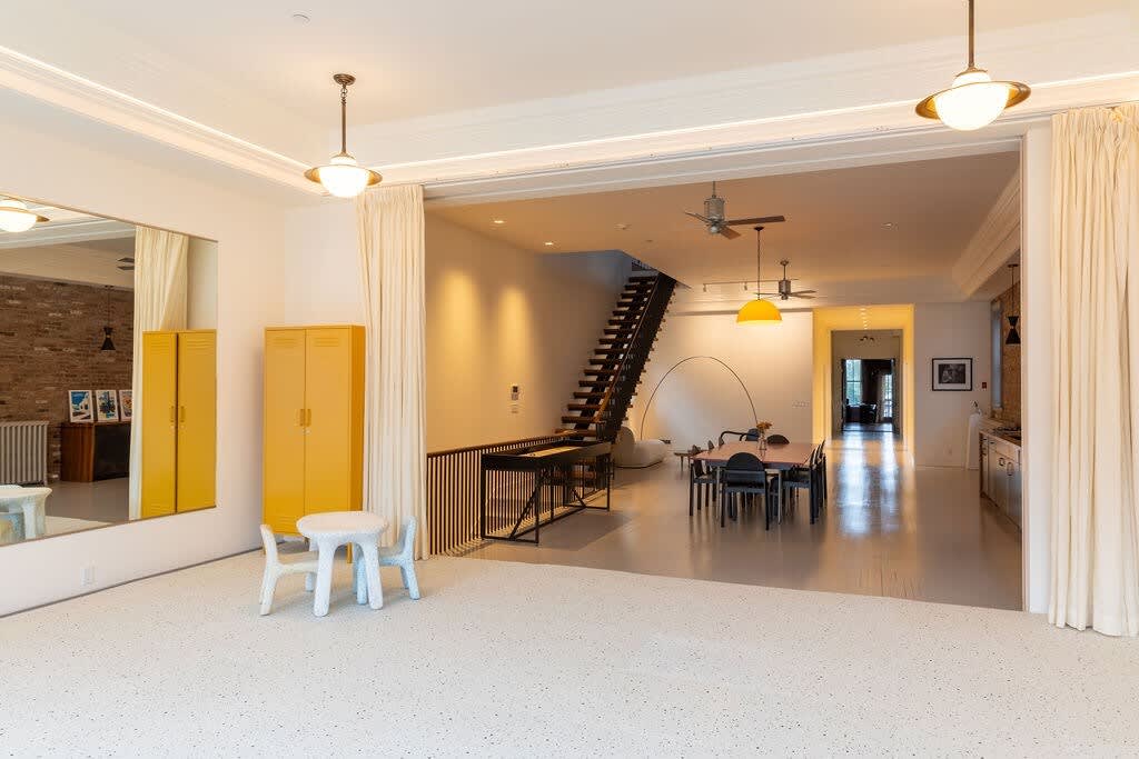Expansive open space with white walls and a speckled floor, featuring a bright yellow locker, a small white children's table, and a view into a lower level with a modern staircase and dining area.