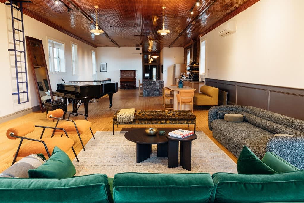 Expansive open-plan loft featuring a living area with a green velvet sofa, a grand piano, and a dining nook, all under a glossy wooden ceiling with exposed beams and track lighting.