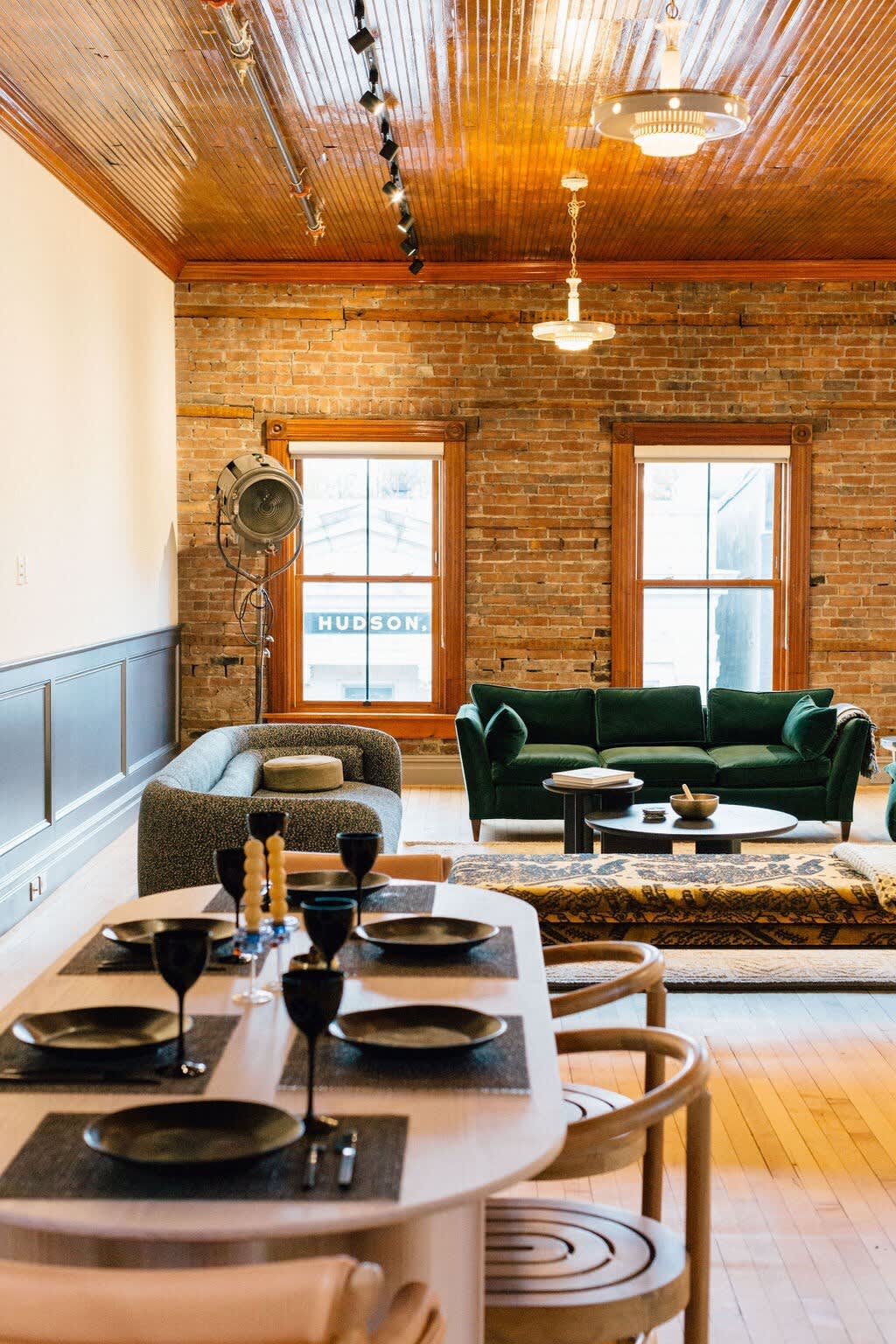 Vibrant open-concept living and dining area with exposed brick walls, a rich wooden plank ceiling, a long dining table, and modern velvet sofas in green and gray.