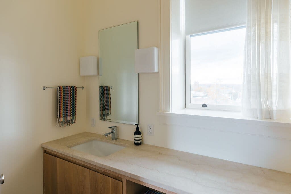 Bright bathroom with a light-colored vanity featuring a rectangular sink, a large mirror, and wall-mounted sconces. A window with sheer curtains offers natural light, and a towel rack with colorful towels is mounted on the wall.