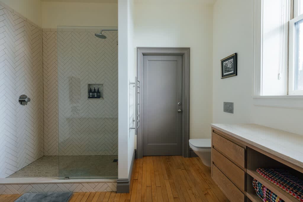 Modern bathroom featuring a walk-in shower with white herringbone tile walls and a glass enclosure, light wood flooring, and a long wooden vanity with a light countertop. A toilet is visible next to the vanity.